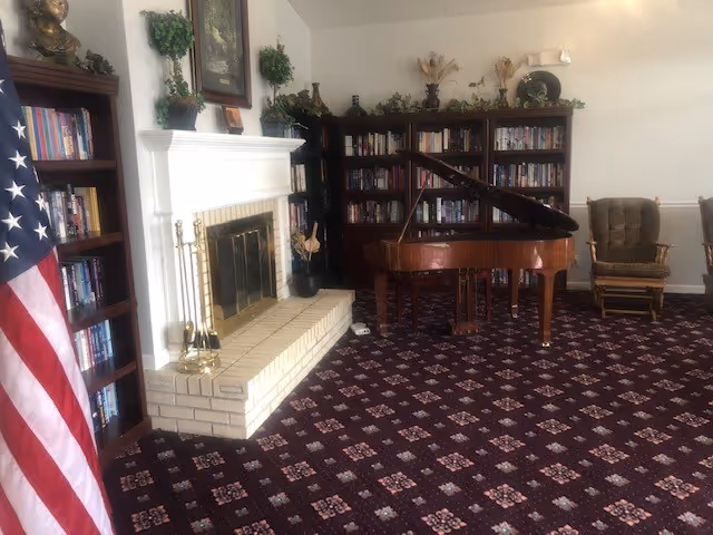 A living room features a brick fireplace, grand piano, bookshelves, flag, and patterned carpet.
