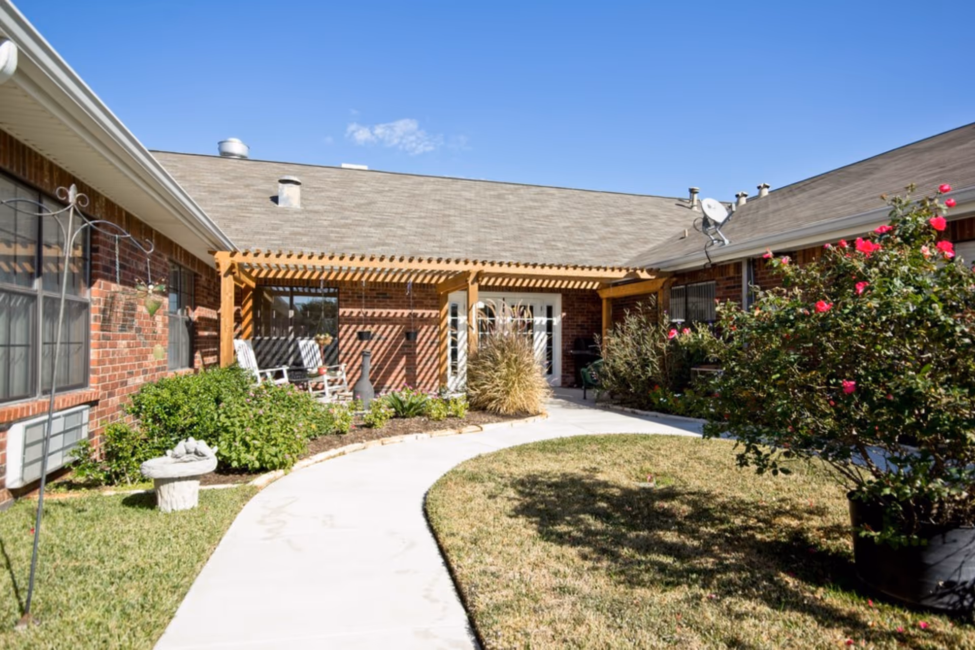 Sunlit courtyard with a curved walkway leading to a pergola-covered entrance on a brick senior living building, with chairs, shrubs, and flowering bushes.