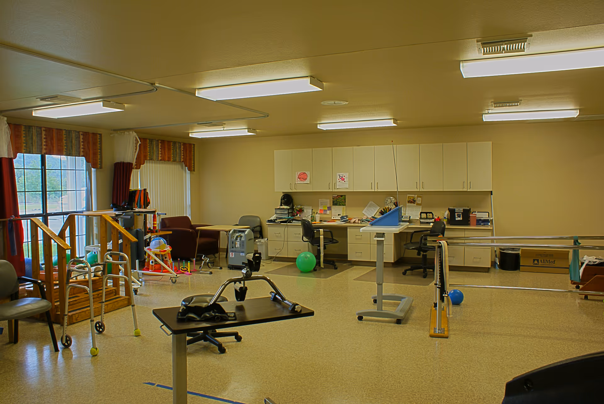 A therapy room with various rehabilitation equipment including parallel bars, a walker, exercise balls, a small wooden staircase, chairs, desks with office supplies, and cabinets along the back wall. The room has large windows with curtains and fluorescent ceiling lights.