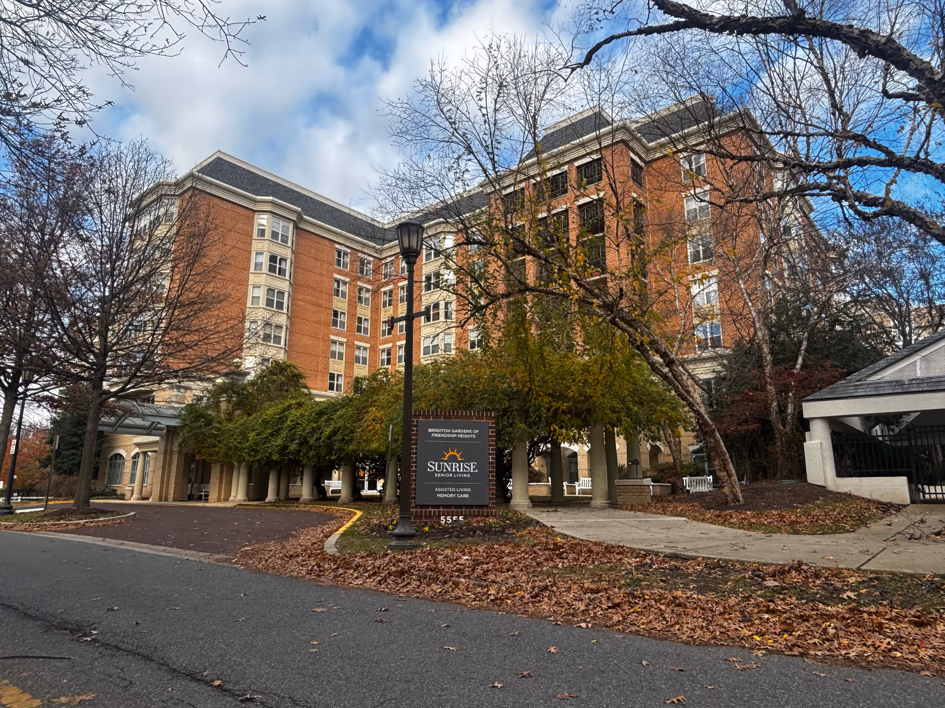 Large multi-story brick senior living building front with a driveway, trees, and a 'Sunrise' sign.