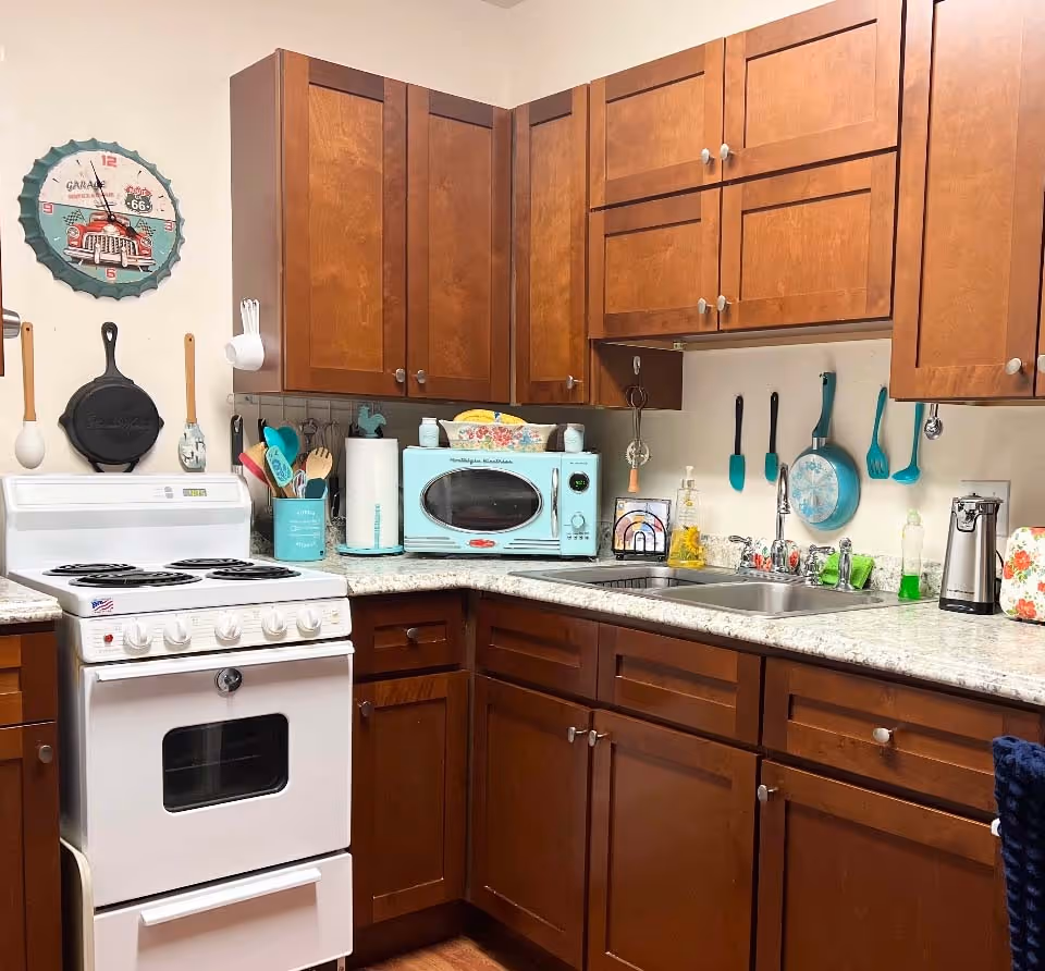 A cozy kitchen corner with wooden cabinets, a white electric stove, a light blue microwave, and various kitchen utensils hanging on the wall. The countertop holds a paper towel holder, soap dispenser, and other kitchen essentials.