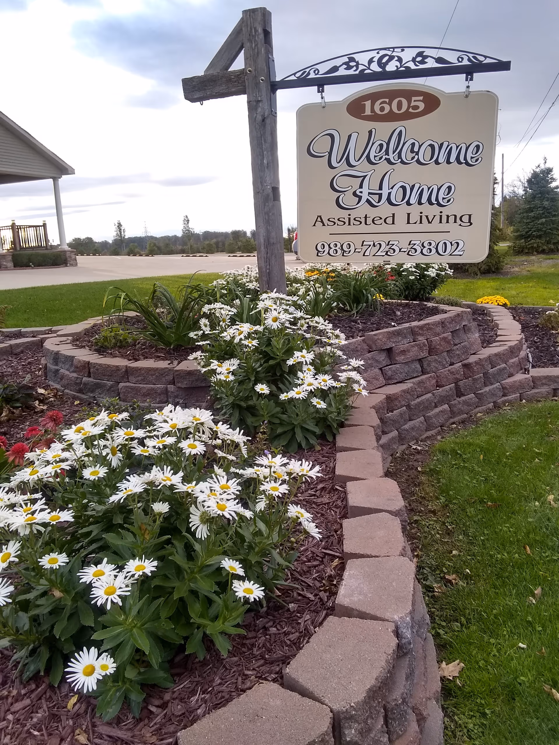 Welcome Home Assisted Living sign with brick-edged flower beds and daisies.