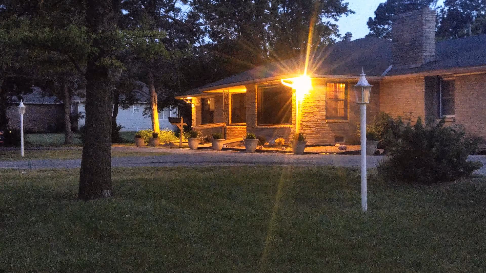 Evening exterior of Residence Begonia Home Plus with lit front porch, lawn, trees, and potted plants.