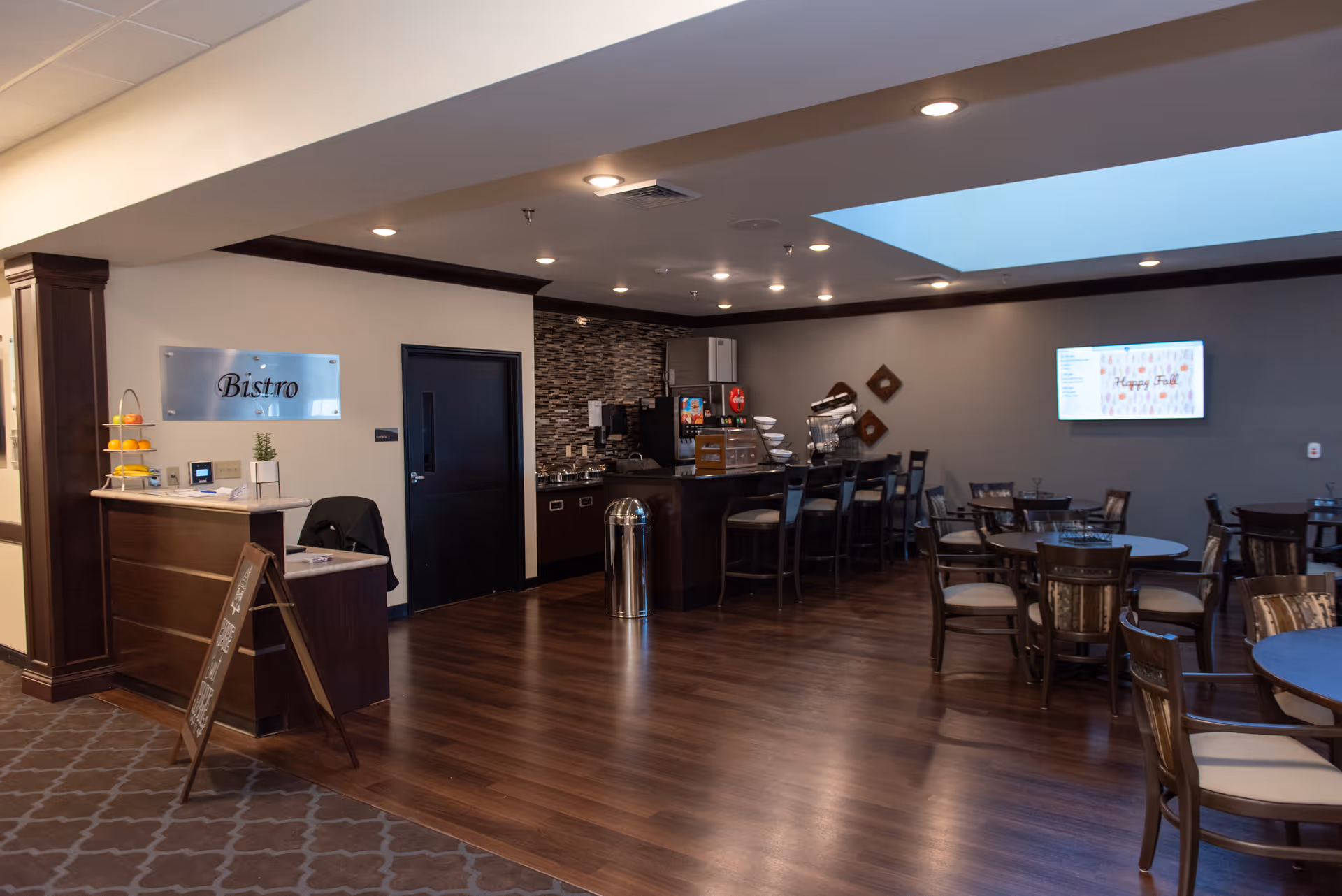 Interior view of a bistro dining area with wooden floors, several round tables with chairs, a counter with bar stools, a drink dispenser, and a sign on the wall that says 'Bistro'. The space is well-lit with recessed ceiling lights and has a modern, cozy atmosphere.