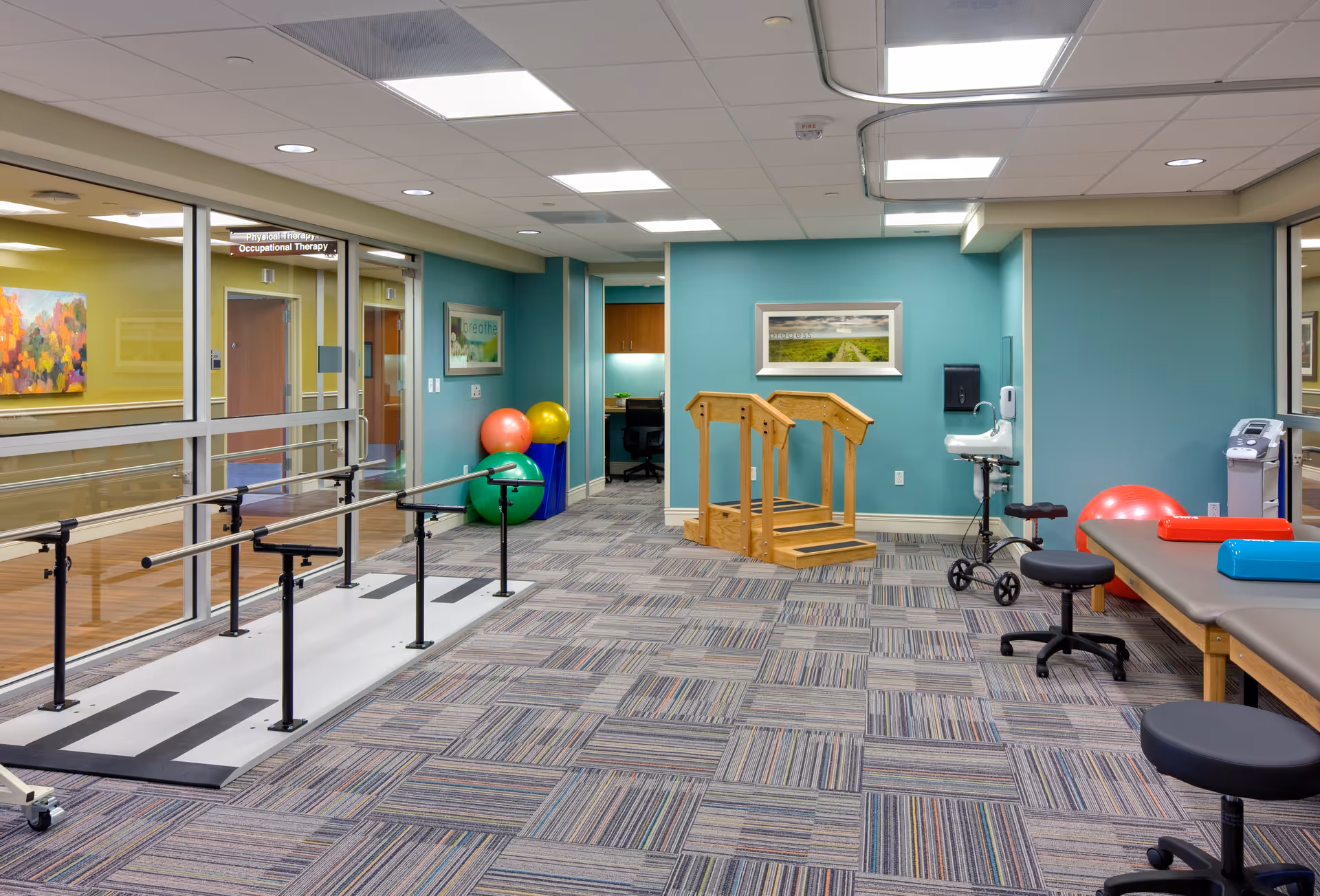 This image features interior senior living therapy gym with parallel bars, exercise balls, and teal walls.