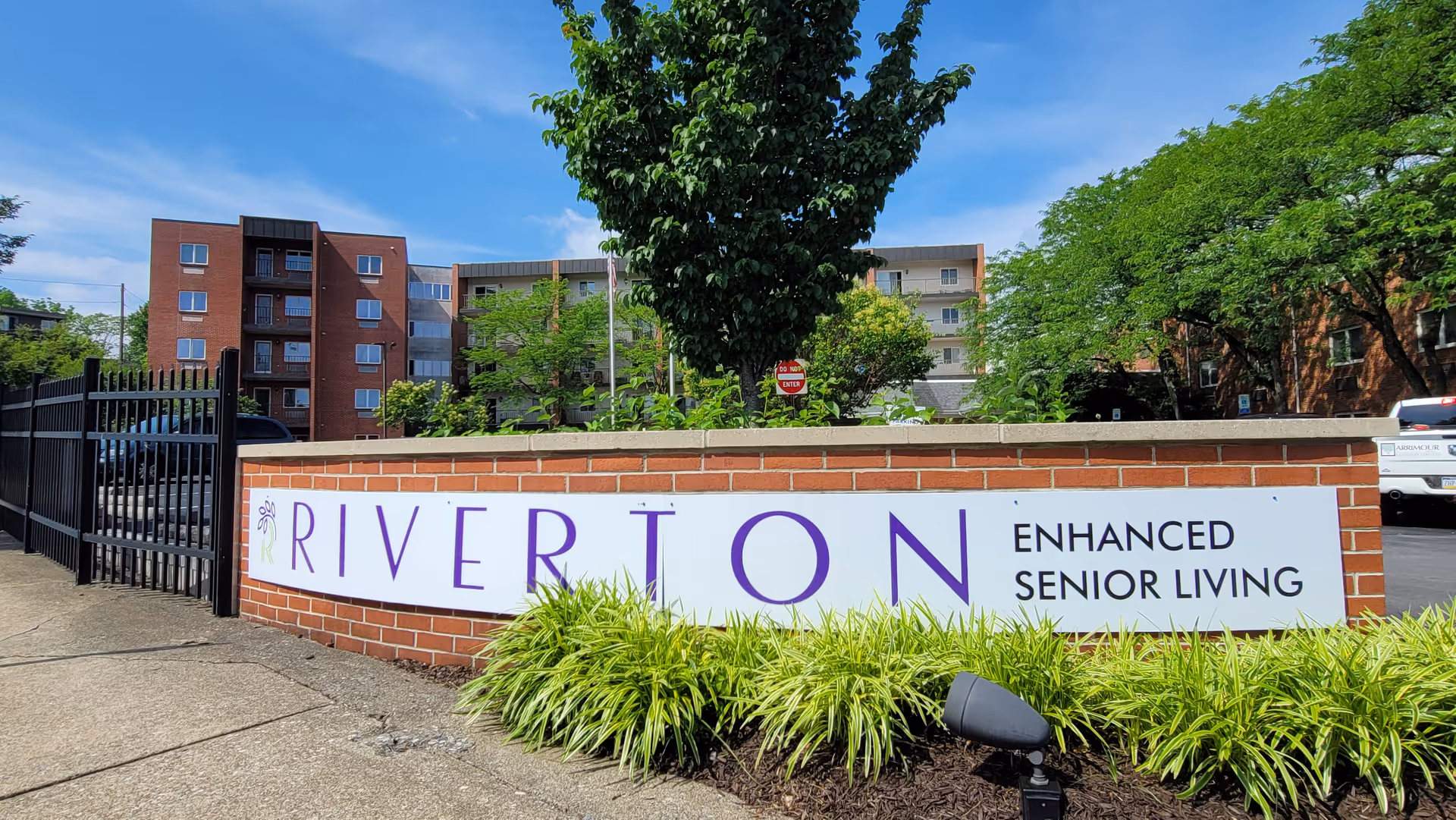 Brick entrance wall with Riverton Enhanced Senior Living sign and surrounding greenery.