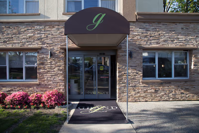 Entrance of a stone-faced building with a brown canopy and glass doors, landscaped with pink flowers.