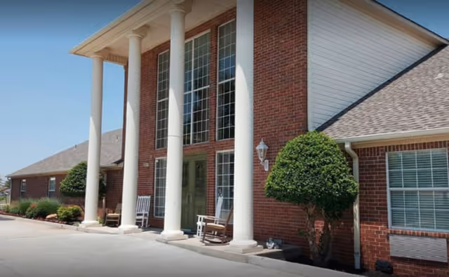 Exterior view of a brick building with tall white columns at the entrance, large windows, two rocking chairs on the porch, and neatly trimmed bushes along the side.