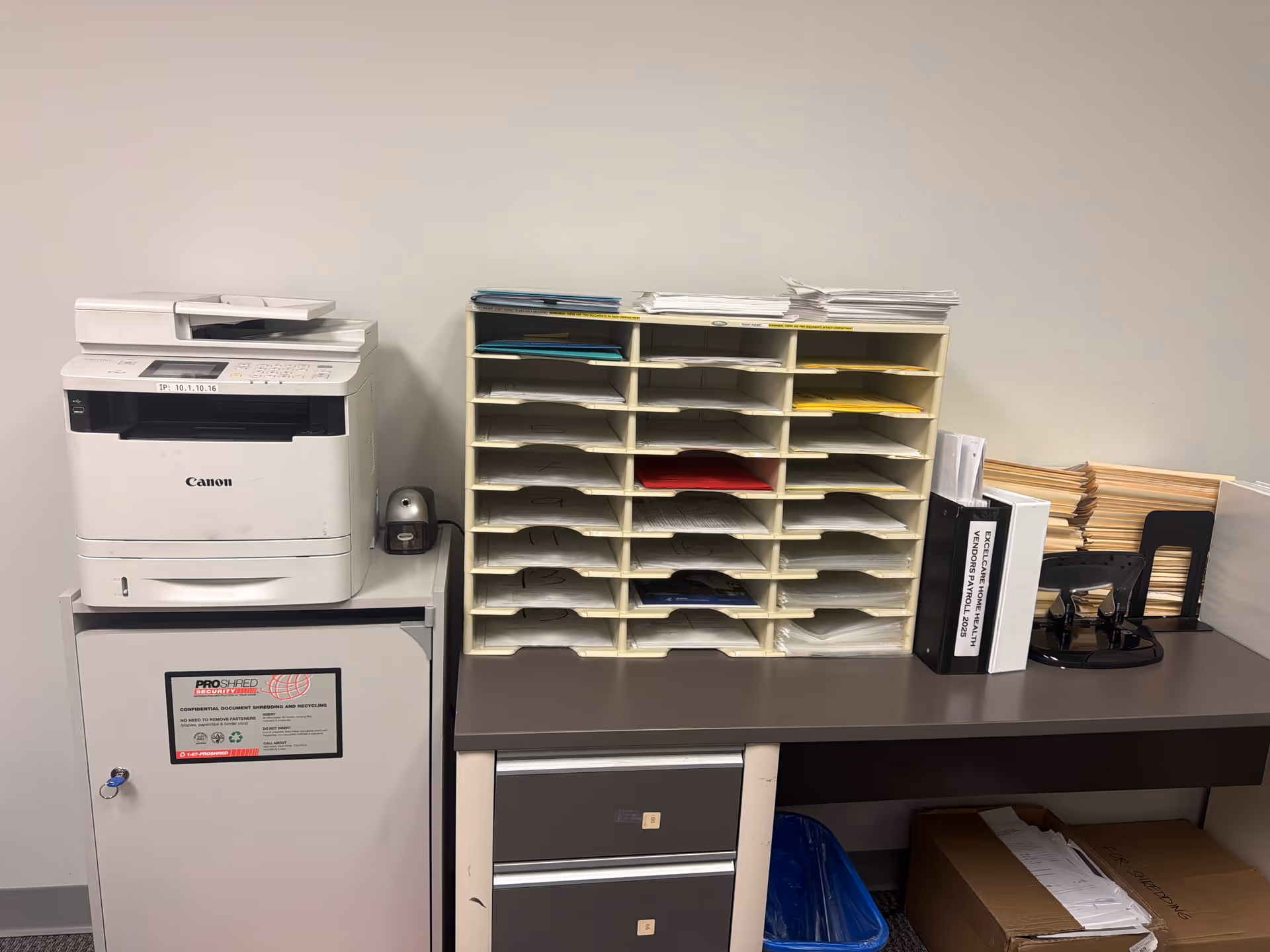 Office workspace with a Canon copier, organized paper trays, folders, and a filing cabinet.