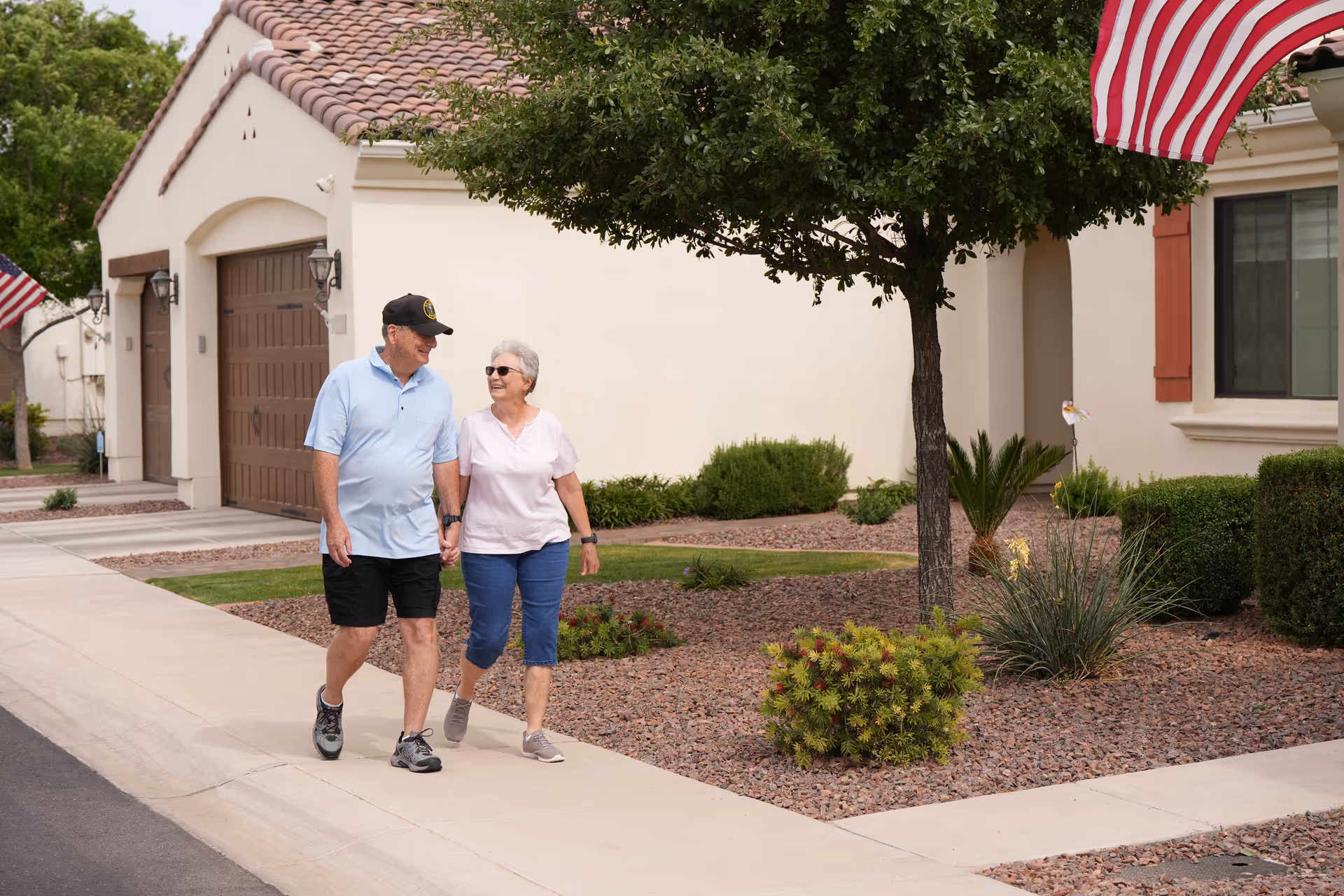An elderly couple holding hands and walking on a sidewalk in a residential area with beige buildings, desert landscaping, and an American flag visible.