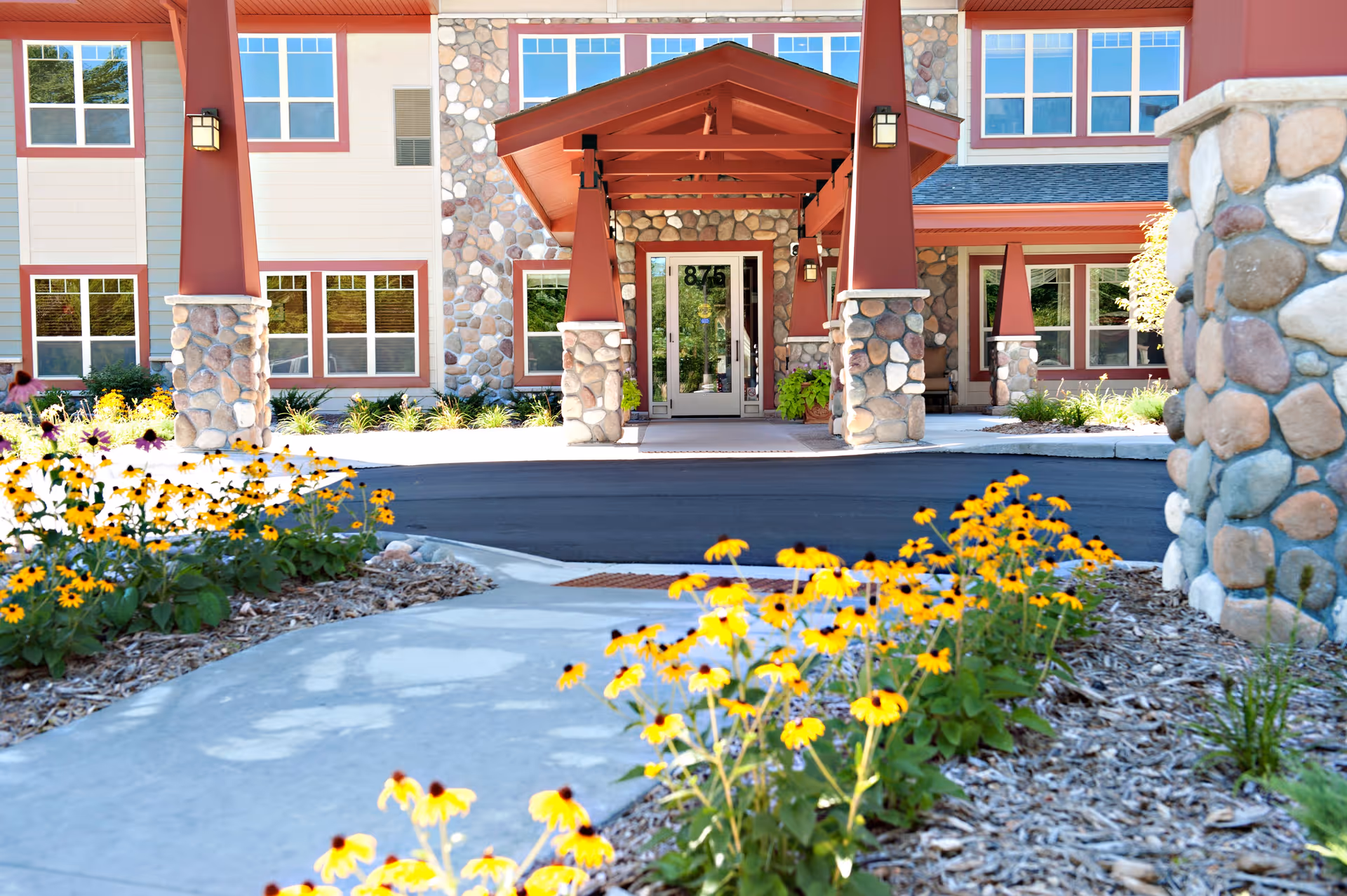 Entrance of Trails of Orono Assisted Living & Memory Care building with stone pillars and a covered walkway. Yellow flowers and landscaping line the concrete path leading to the glass door entrance.