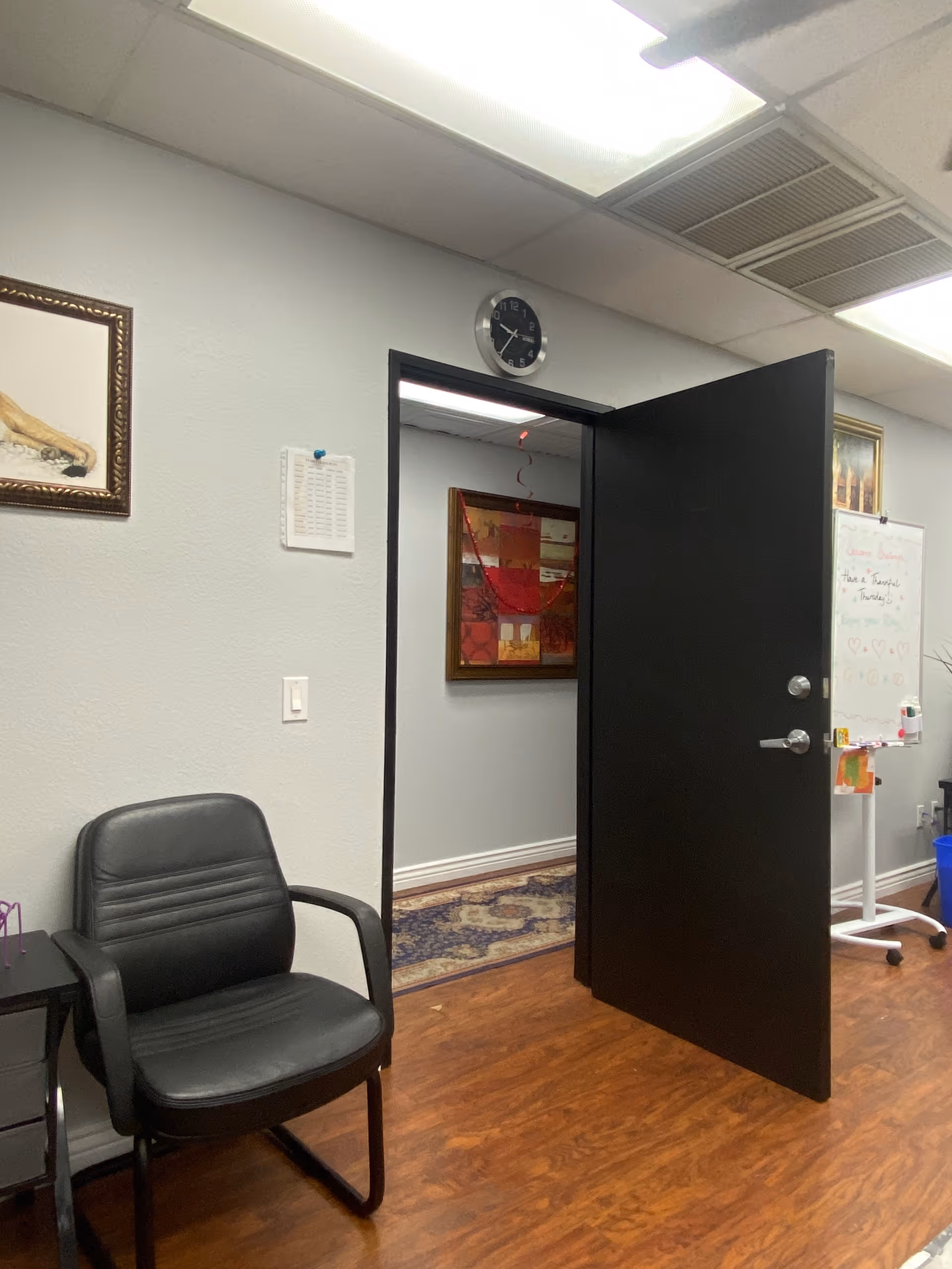 Interior office-like room with black chair, open doorway, wall clock, and whiteboard.