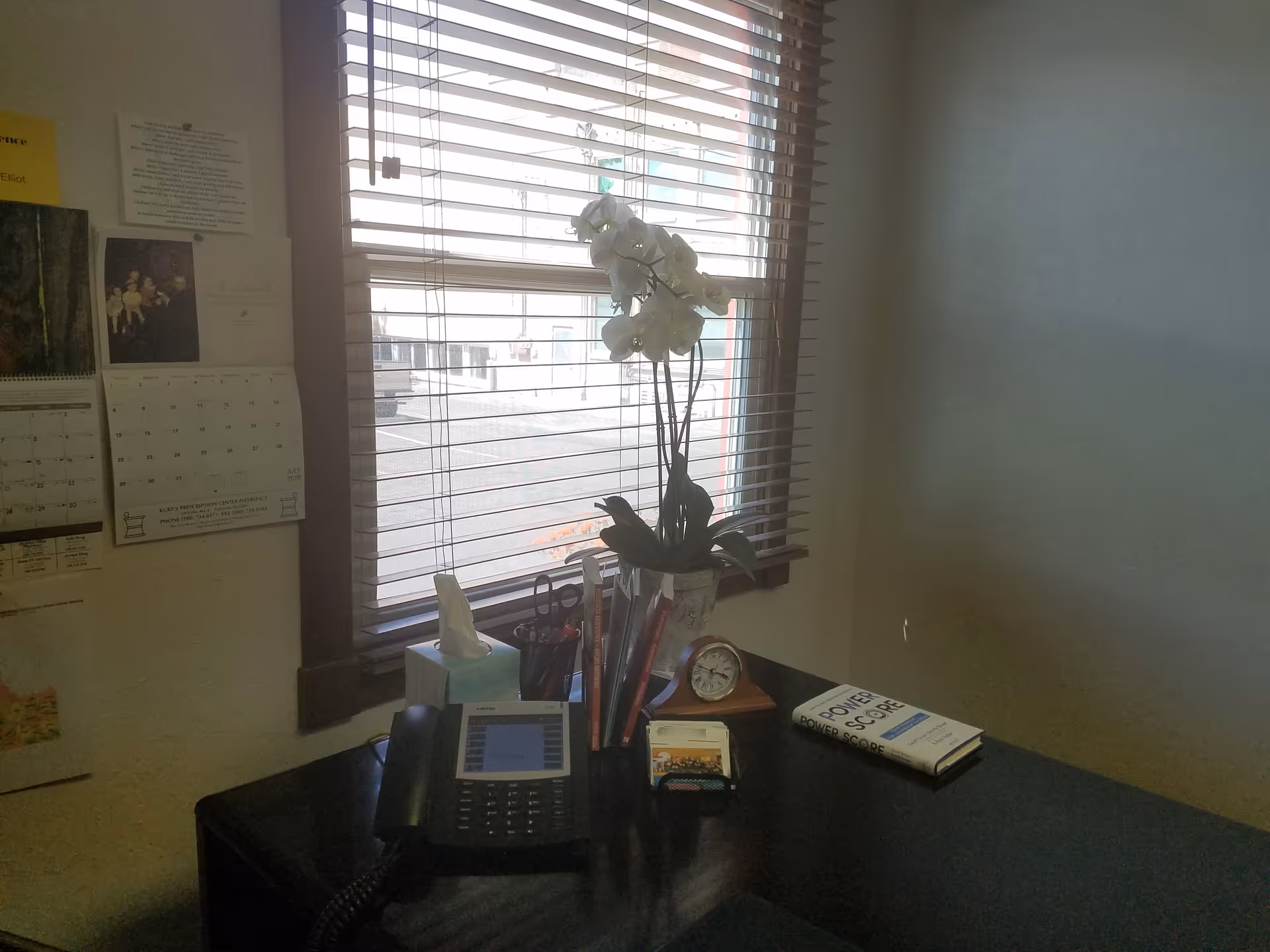 Office desk with landline phone, clock, tissues, and a potted orchid by window blinds.