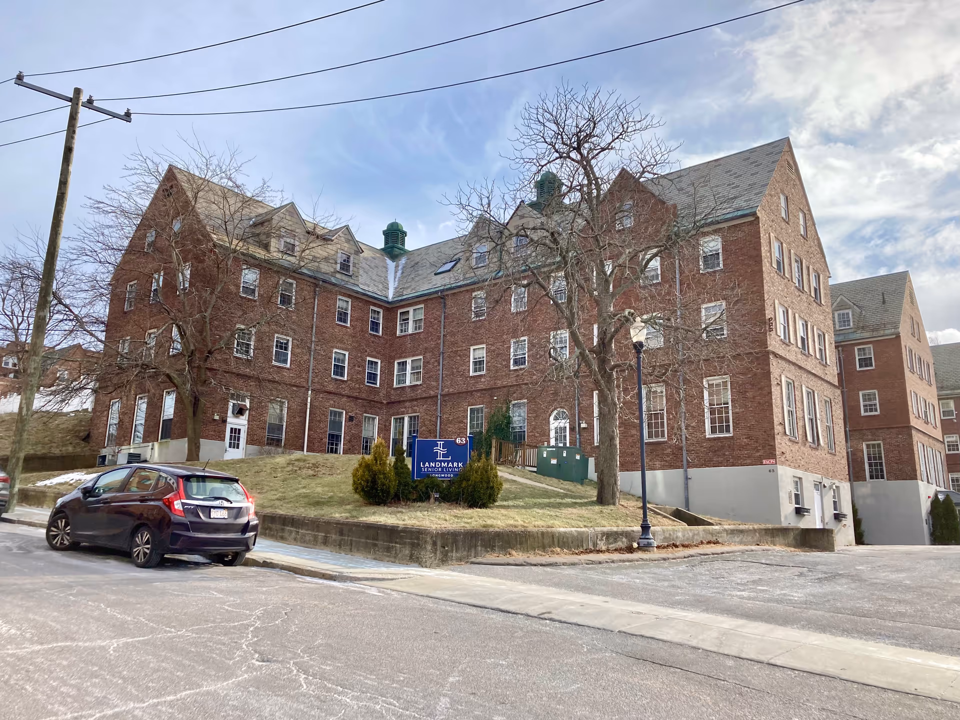 Large brick senior living building on a slight hill with a 'Landmark Senior Living' sign, leafless trees, and a parked car in front.