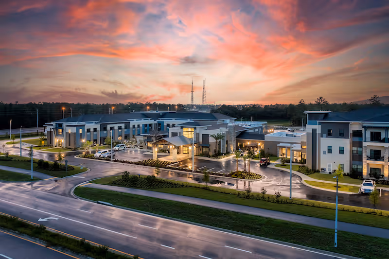 Aerial exterior view of Oak Hill Senior Living at sunset showing the illuminated main entrance, parking lot, and surrounding roads.