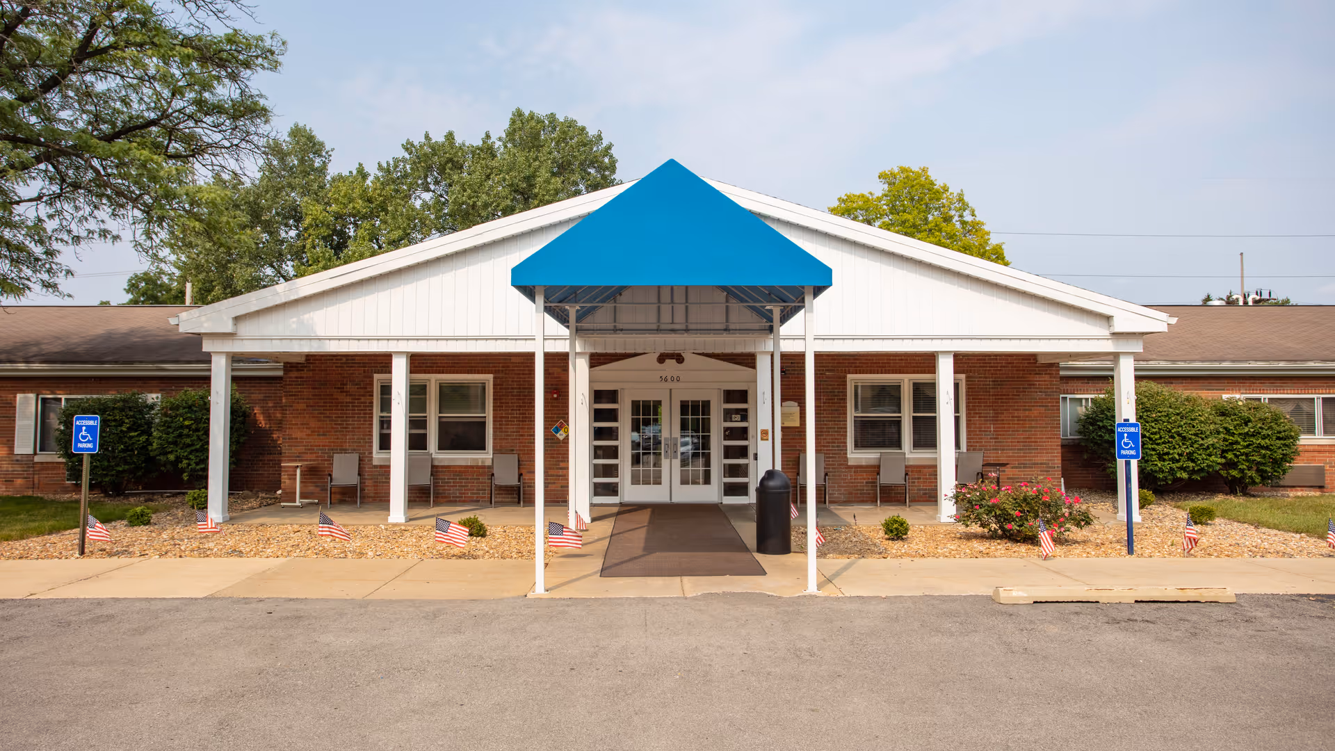 Front exterior view of a single-story brick building with a white gabled roof and a blue awning over the entrance. There are two handicap parking signs on either side of the entrance, small American flags planted in the landscaping, and some chairs placed near the windows. Trees and bushes surround the building.