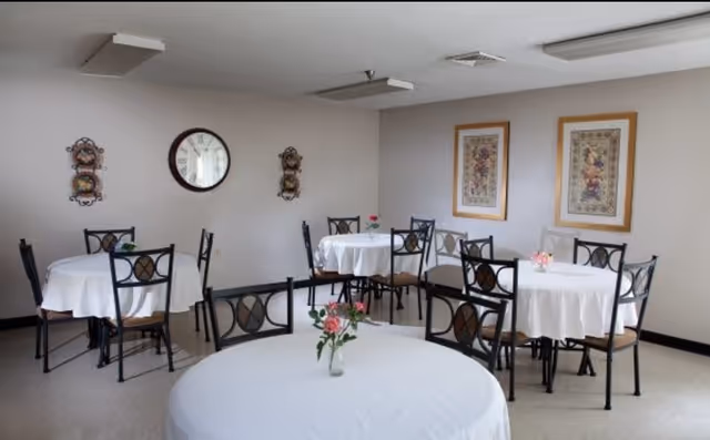 A small dining room with round tables covered in white tablecloths, each table having a small vase with flowers. The room has several black metal chairs with cushioned seats. On the walls, there is a round clock and framed floral artwork.