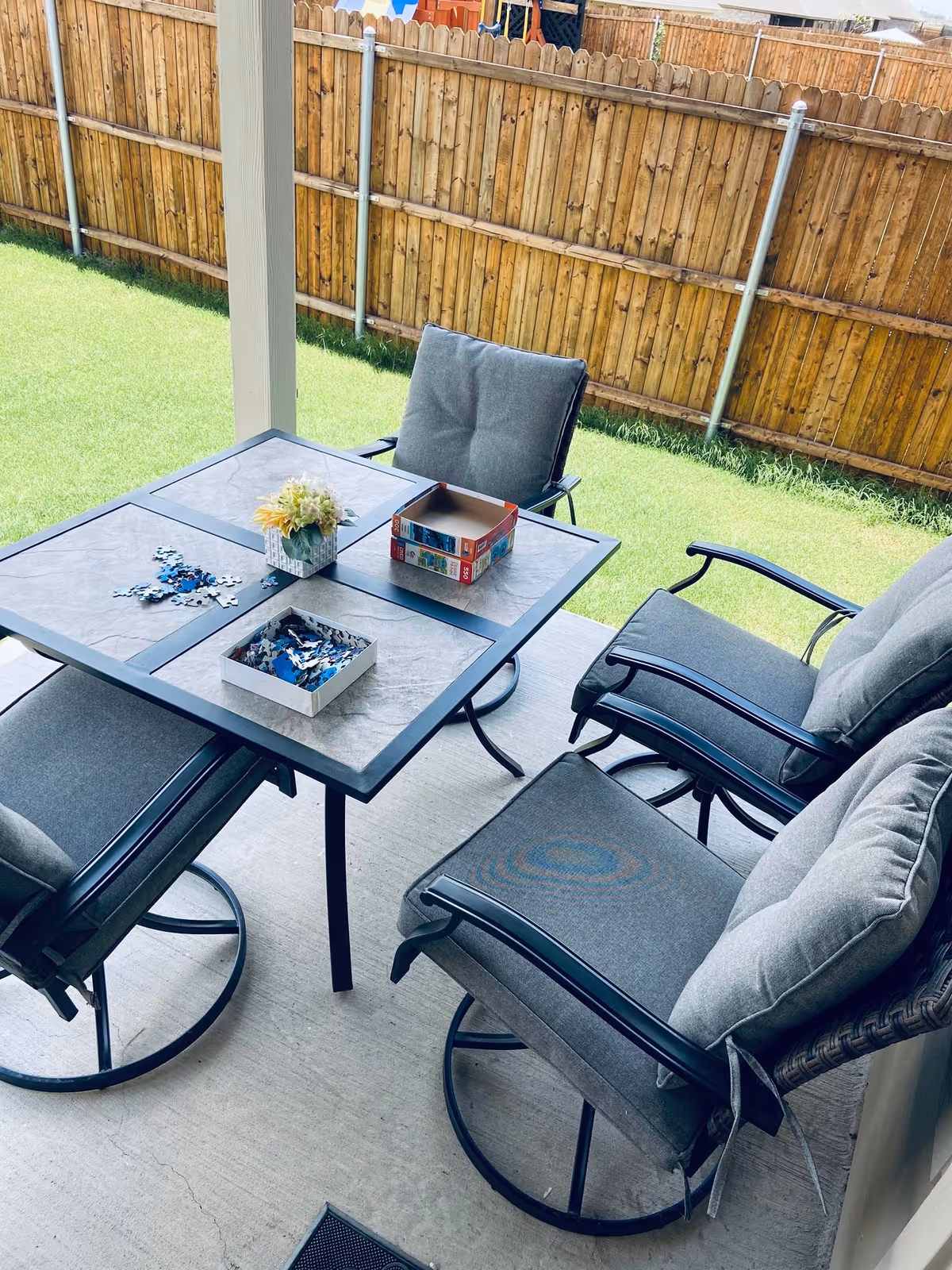 Outdoor patio area with cushioned chairs, a table puzzle, and a fenced grassy backyard.