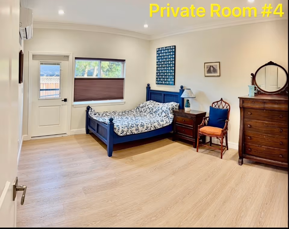 A private bedroom with a blue bed frame, white walls, window with blinds, and wooden dresser.