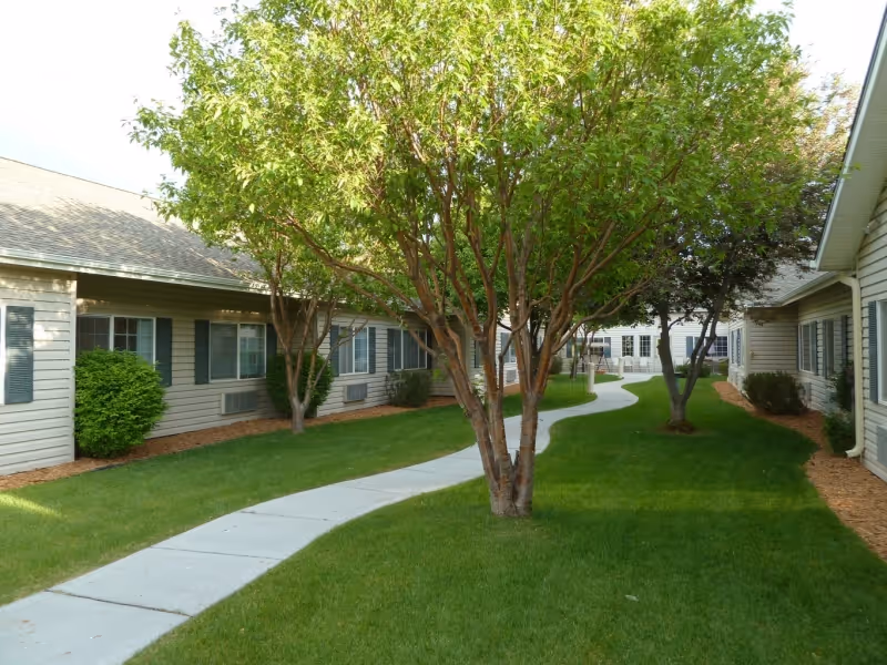 A winding concrete pathway runs through a grassy courtyard area with several trees and shrubs, bordered by single-story buildings with beige siding and green shutters.