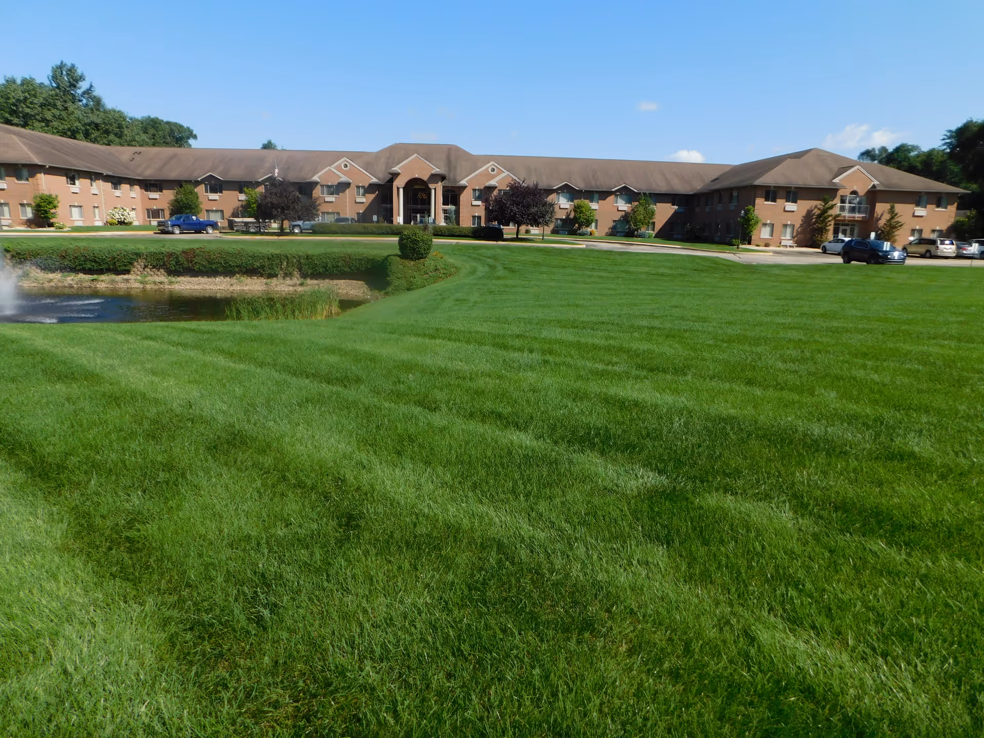 Wide view of a long brick senior living building set behind a large manicured lawn and a small pond with a fountain.