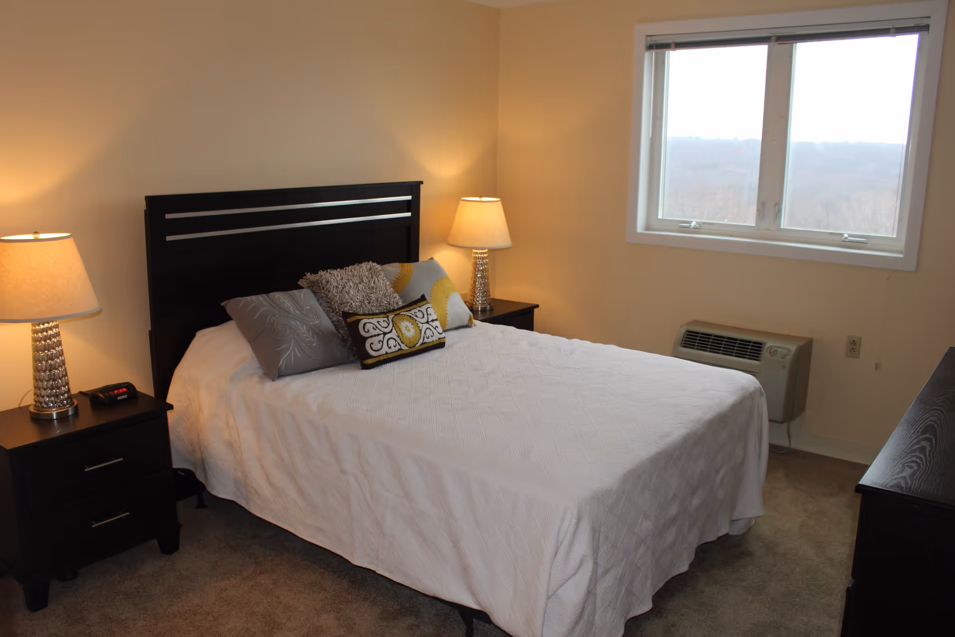 A bedroom with a neatly made bed covered with a white quilt and several decorative pillows. There are two dark wooden nightstands on either side of the bed, each with a lamp. A window on the right side shows an outdoor view, and there is a wall-mounted air conditioning unit below the window. The room has beige walls and carpeted flooring.