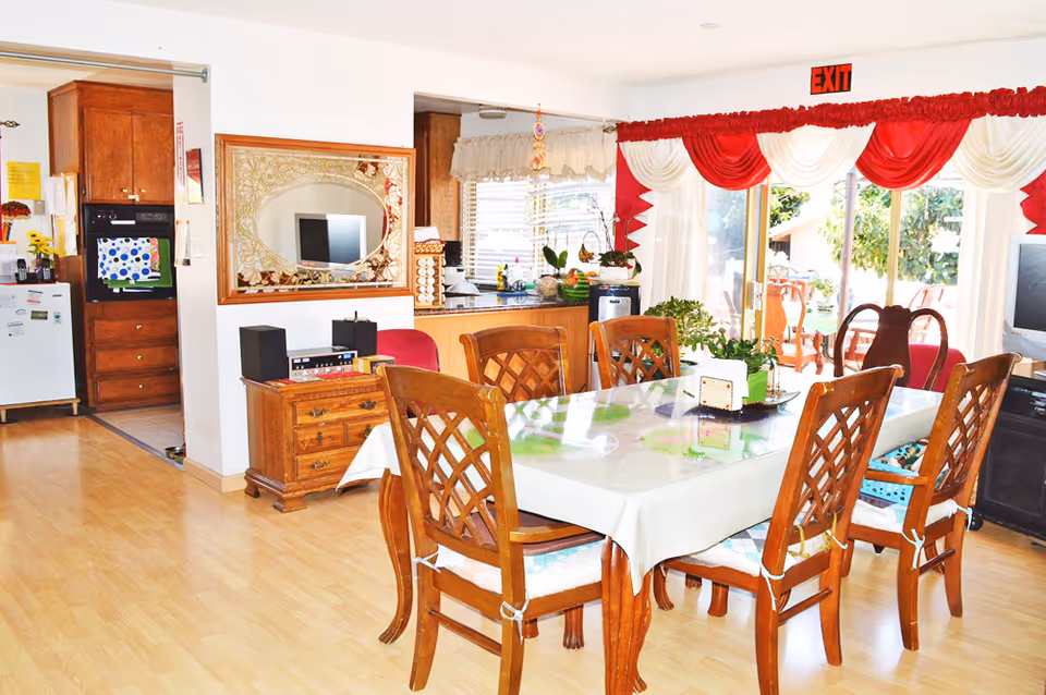 A bright dining area with a wooden dining table covered with a white tablecloth and surrounded by six wooden chairs with cushions. The room has light wooden flooring and a large window with red and white drapes, leading to an outdoor patio with chairs and greenery visible outside. To the left, there is a kitchen area with wooden cabinets, a refrigerator, and an oven. A decorative mirror hangs on the wall above a wooden side table with electronic devices on it. An exit sign is visible above the window.