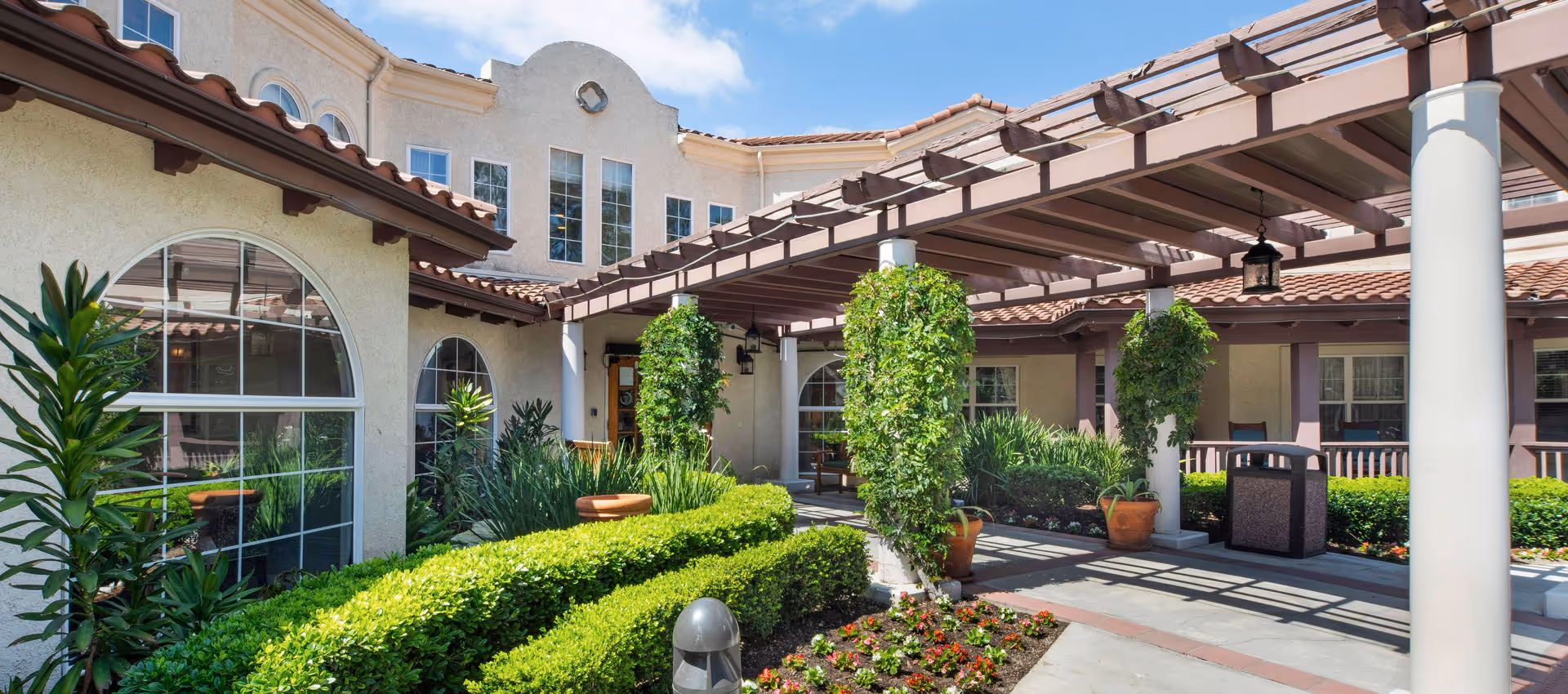 Exterior view of Sunrise Of Mission Viejo facility showing a covered walkway with columns wrapped in greenery, manicured bushes, potted plants, and arched windows on a sunny day with blue sky and some clouds.