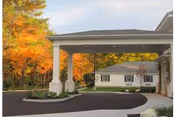 A covered driveway entrance with white columns leads to a senior living facility amid autumn trees.