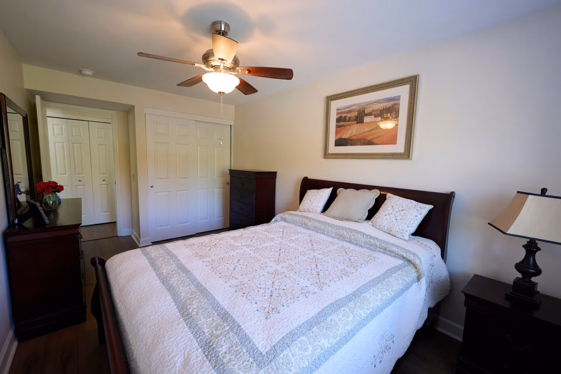 A neatly made bedroom with a king-size bed, dresser, and ceiling fan in soft lighting.