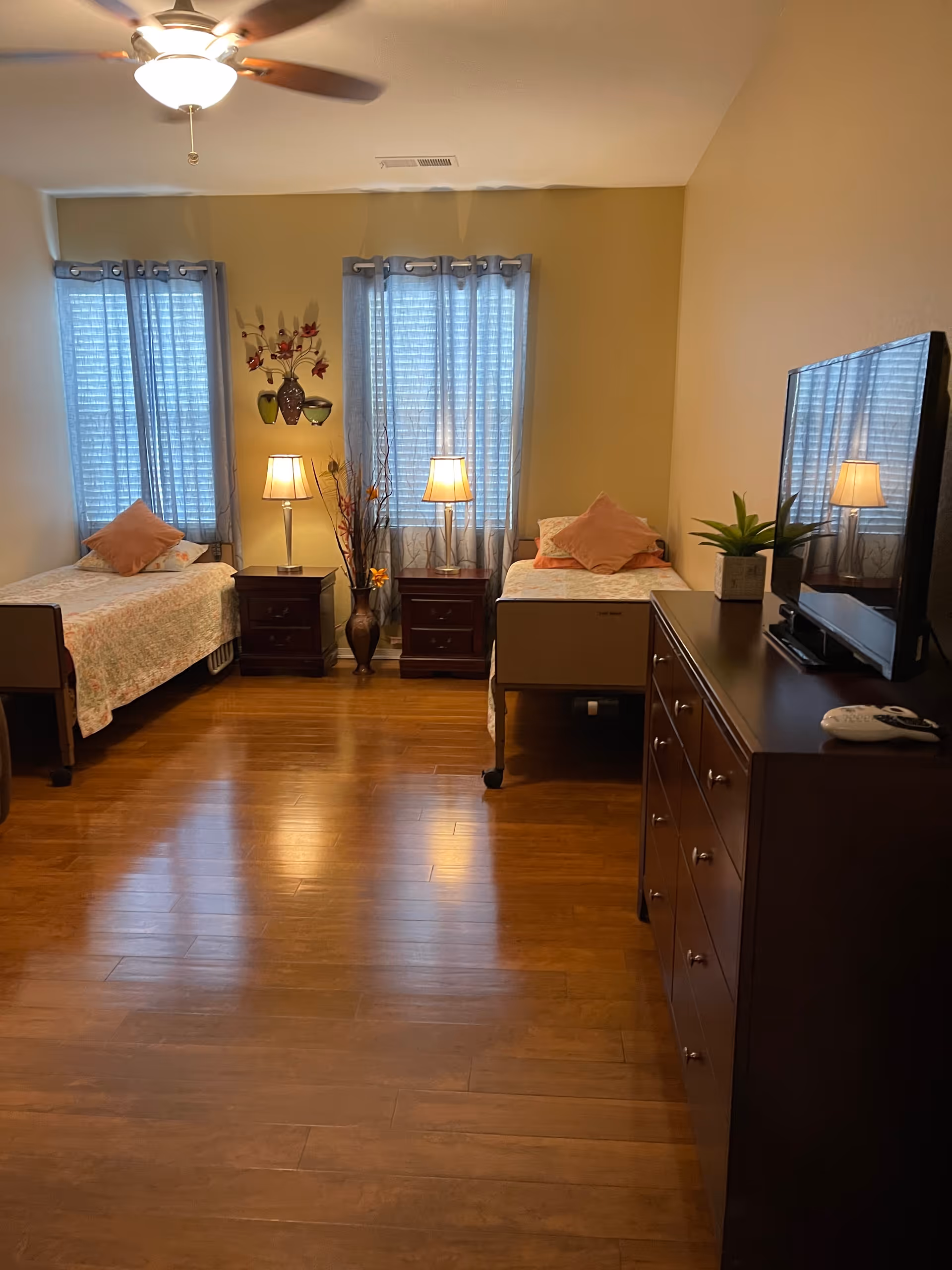 A bedroom with two single beds placed against opposite walls, each with a pillow and floral bedspread. Between the beds are two nightstands with lamps and a decorative vase with flowers. Two windows with blue curtains are behind the beds. On the right side, there is a dark wooden dresser with a flat-screen TV and a small potted plant on top. The room has wooden flooring and a ceiling fan with a light fixture.