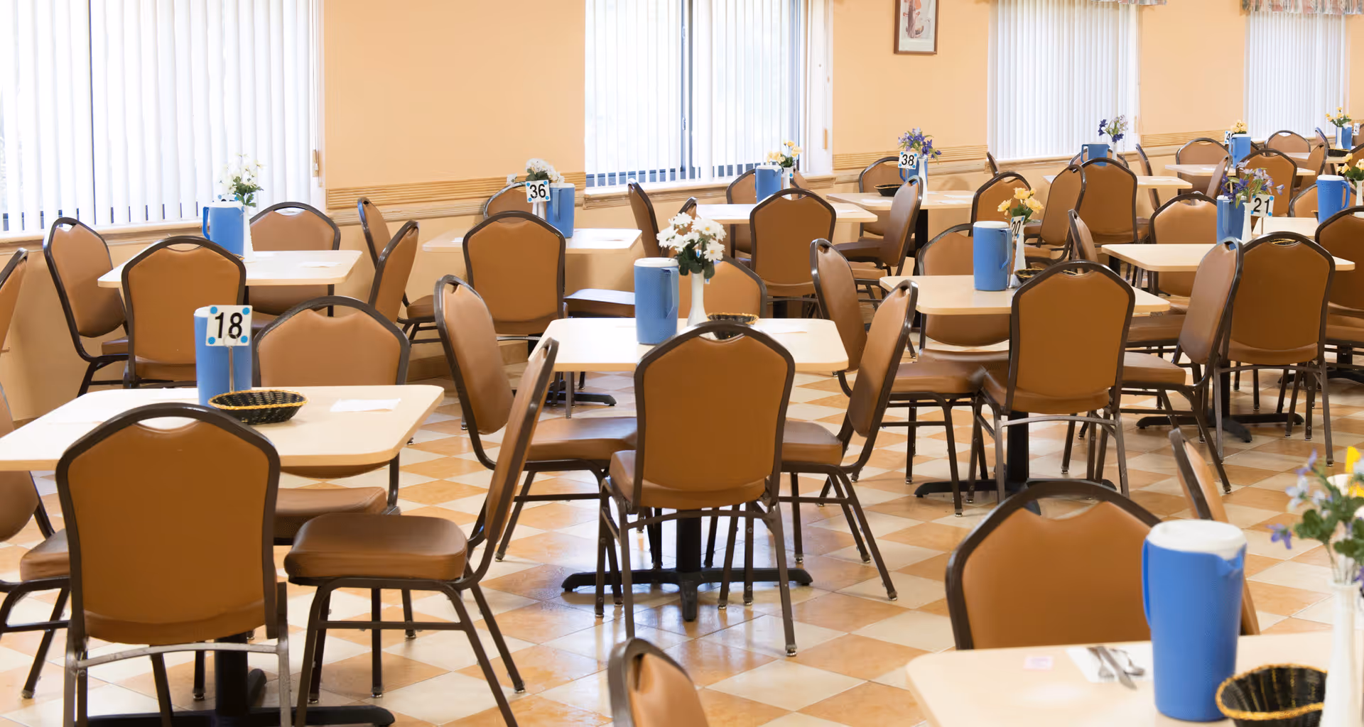 Spacious dining room with multiple tables and brown chairs, each table set with blue pitchers and small flower centerpieces.