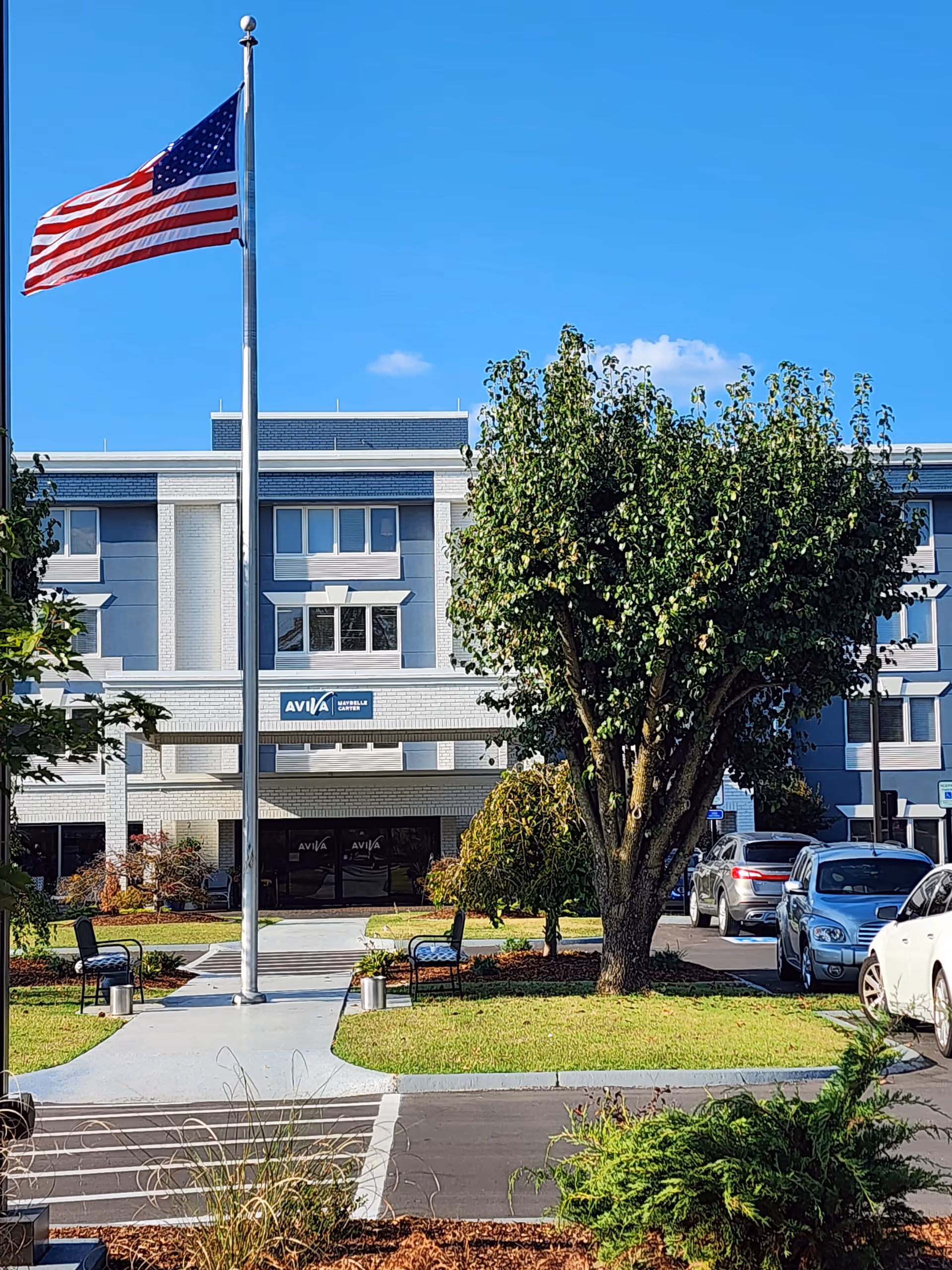 Exterior view of the AVIVA Maybelle Carter senior living facility with a large American flag on a flagpole in front, a tree, benches, parked cars, and a clear blue sky.