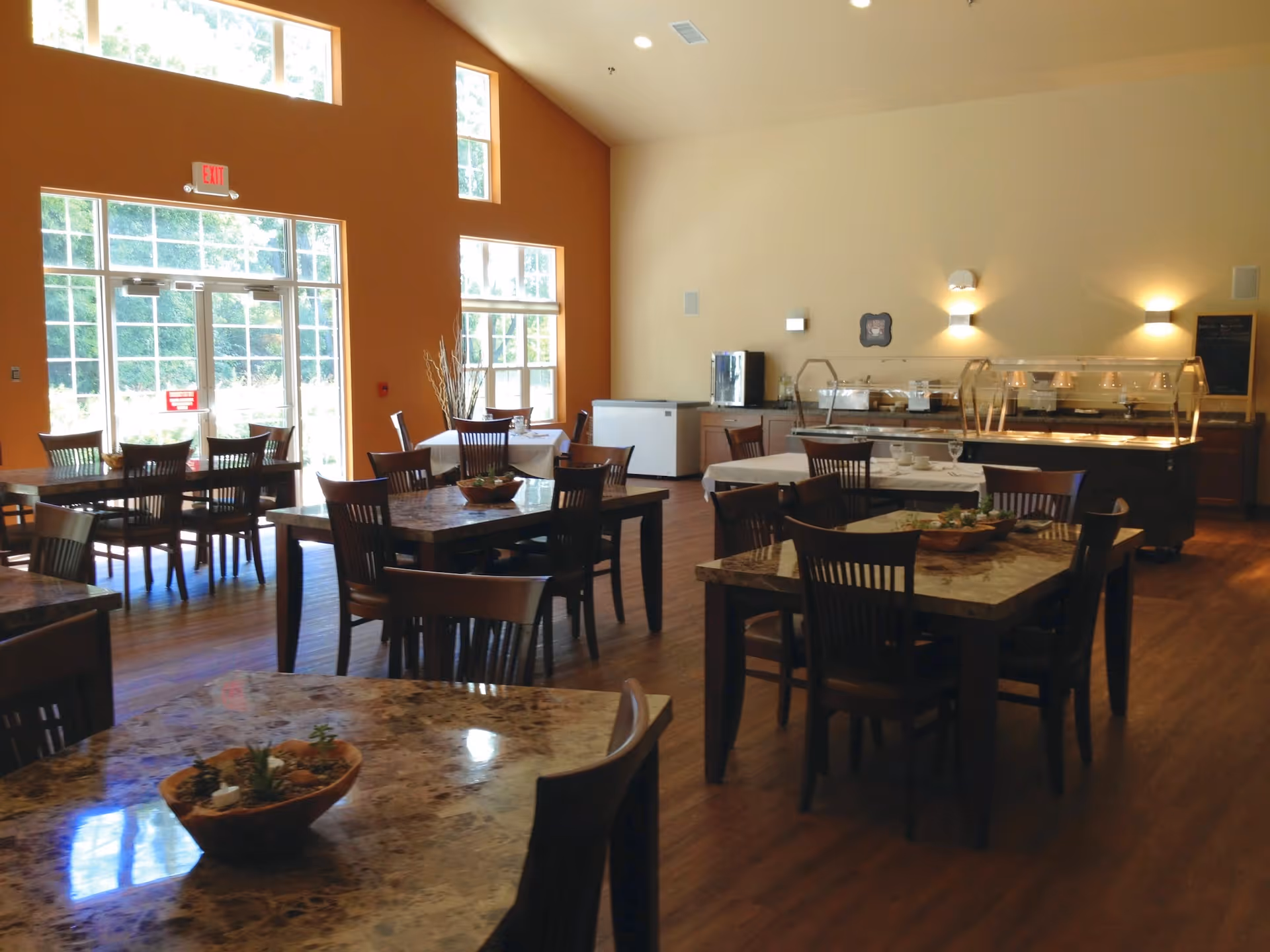 Sunlit dining room with multiple marble-top tables and wooden chairs and a buffet serving station along the back wall.