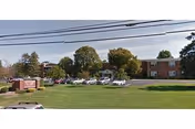 Grassy foreground with parked cars, trees, and a brick building in the distance.