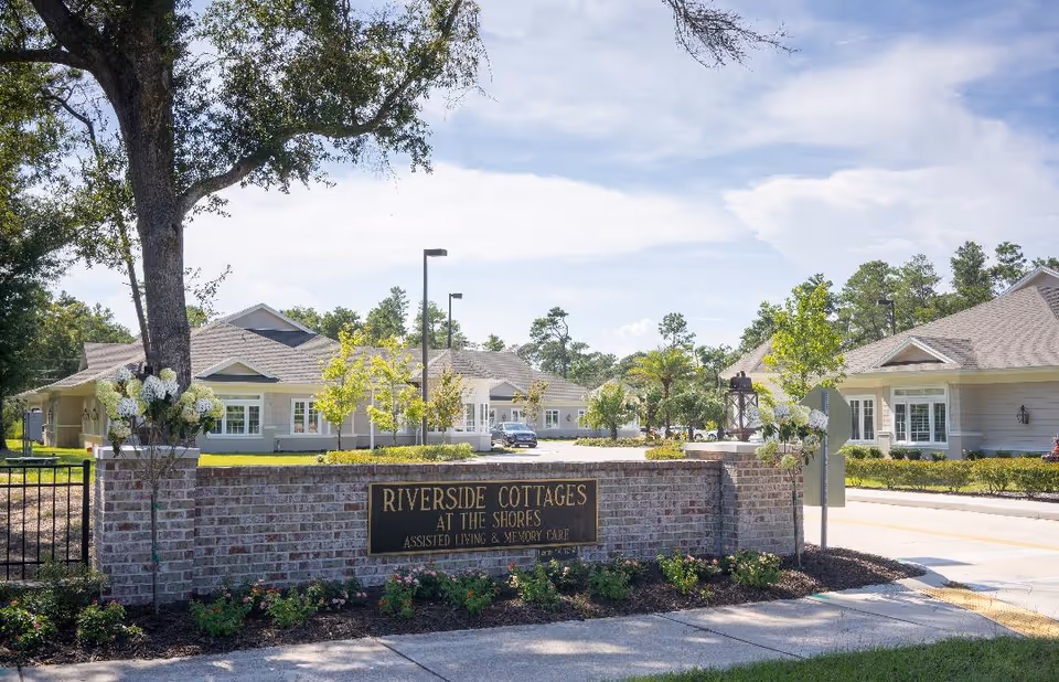 Entrance to Riverside Cottages at the Shores assisted living and memory care facility with a brick sign, landscaped flowers, trees, and single-story buildings in the background under a partly cloudy sky.