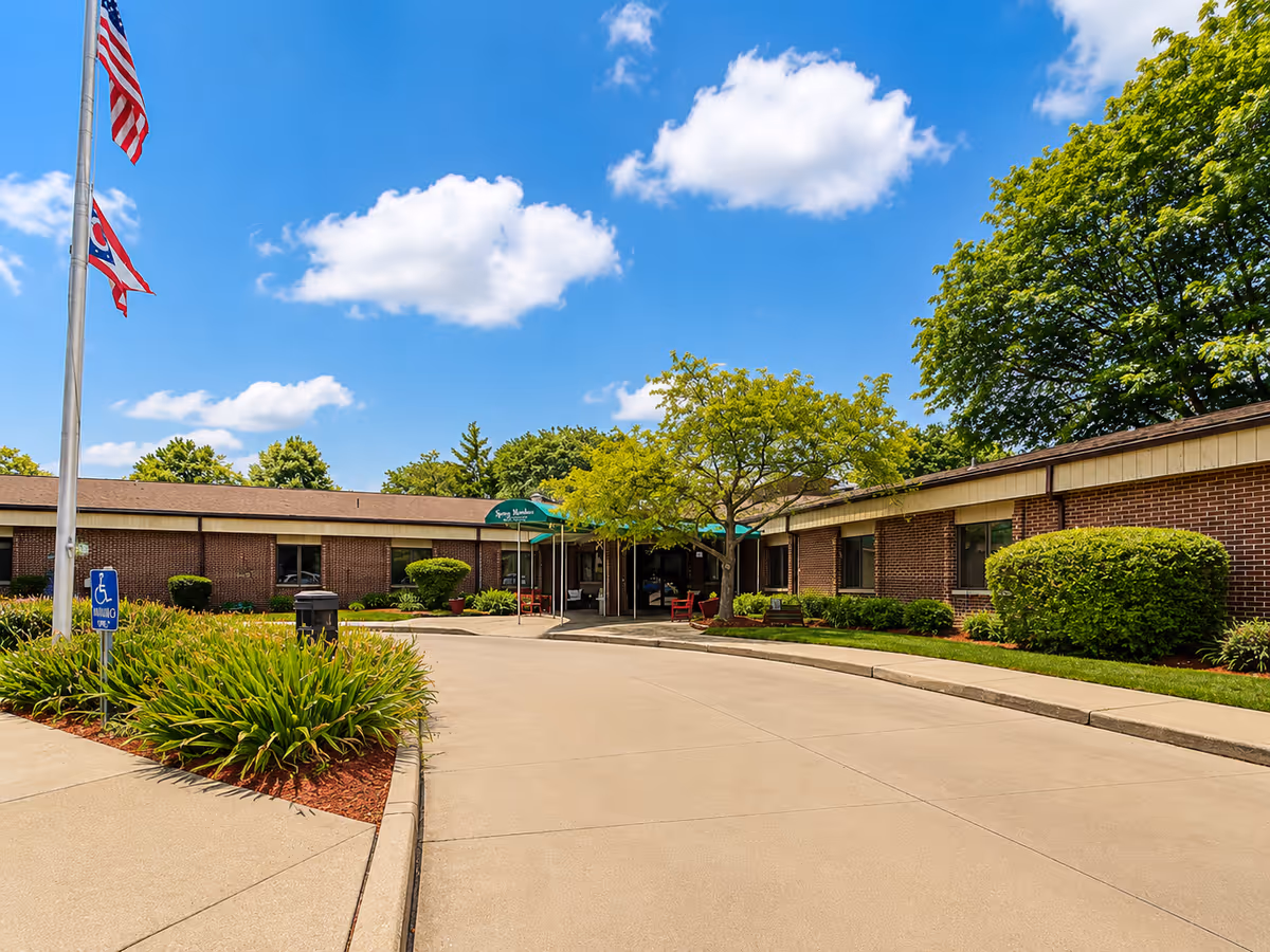 Exterior view of a brick senior living facility with flags, trees, and a curved driveway.