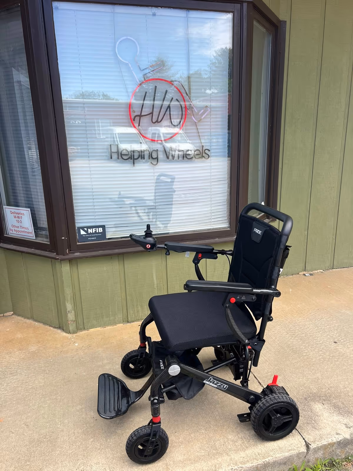 Wheelchair parked outside a building with Helping Wheels Inc sign in window.