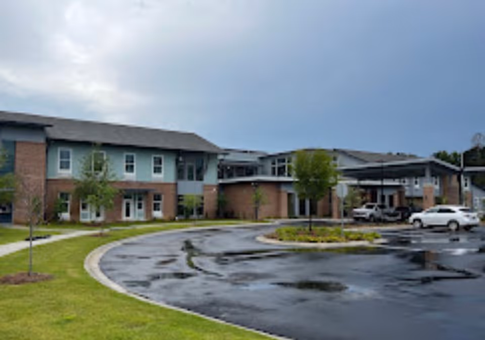 Senior living facility building exterior with driveway, parking, and landscaped grounds.