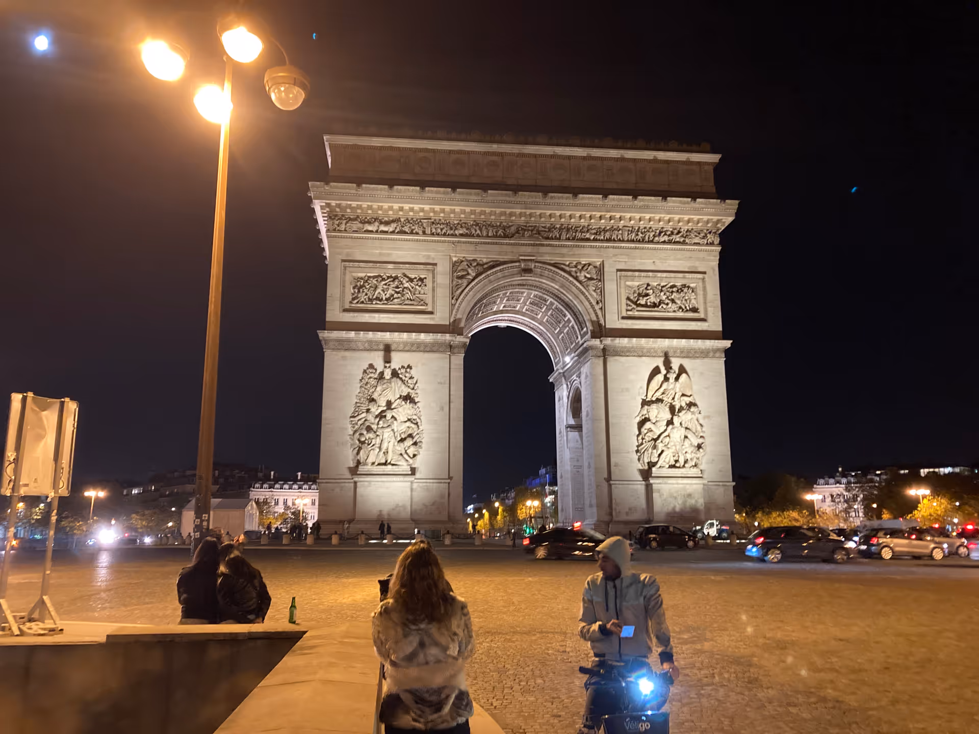 Nighttime view of the Arc de Triomphe with pedestrians and illuminated streetlights.