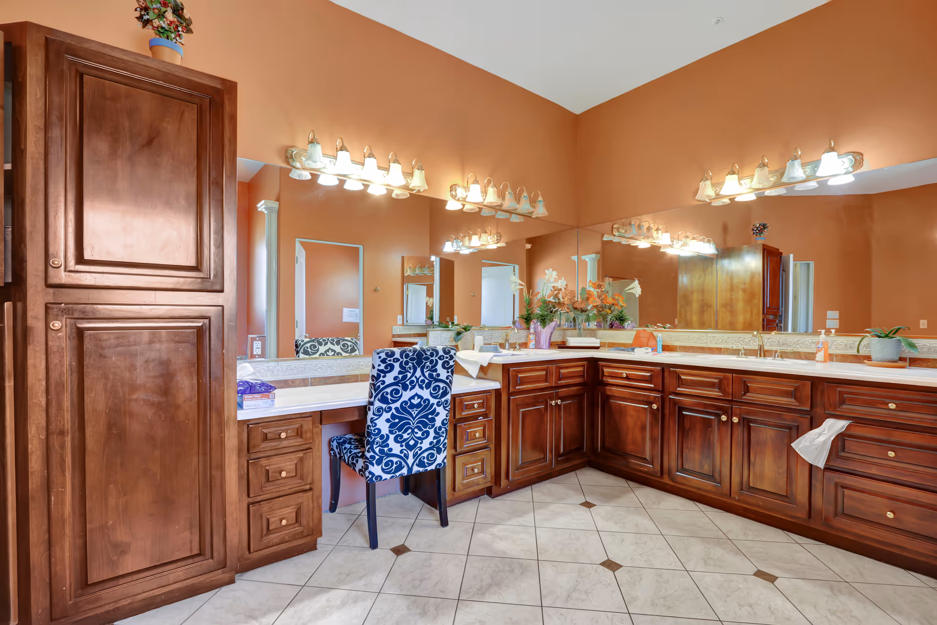 Spacious bathroom vanity area with large mirrors, wooden cabinets, double sinks, and a patterned chair.