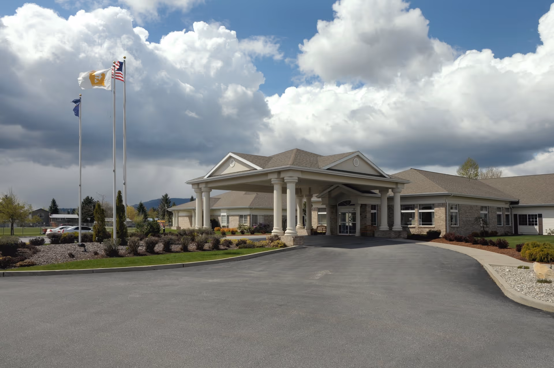 Exterior view of a single-story senior living facility building with a covered entrance supported by columns. There are three flagpoles with flags flying in front of the building, a paved driveway, landscaped bushes, and a partly cloudy sky overhead.