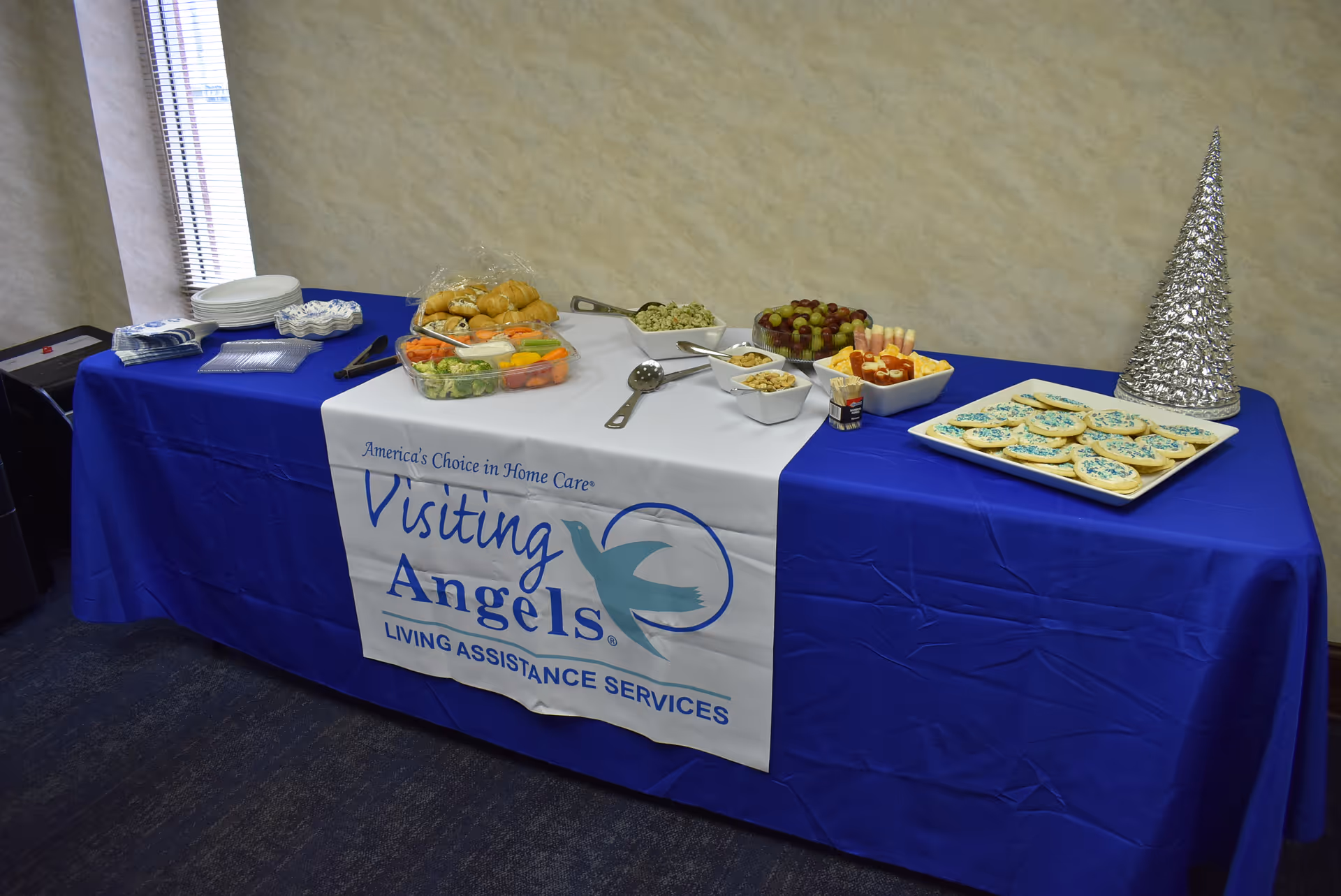 Buffet table with assorted snacks and a Visiting Angels banner on a blue tablecloth.