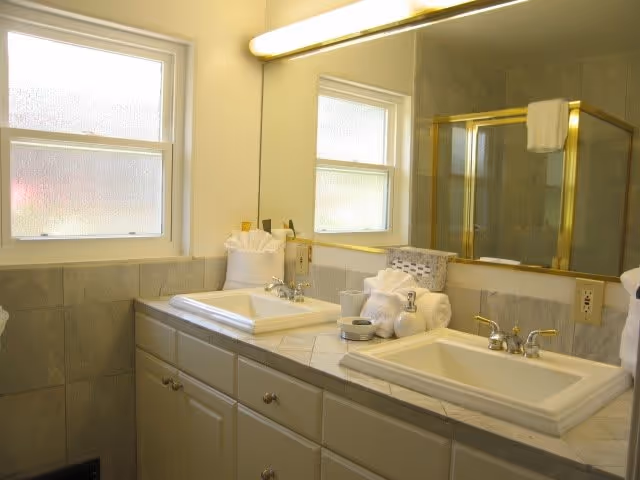A bathroom with two white sinks set in a tiled countertop. Above the sinks is a large mirror with a light fixture. There is a frosted window on the left wall and a glass shower door with gold trim reflected in the mirror. Towels and toiletries are neatly arranged on the countertop.