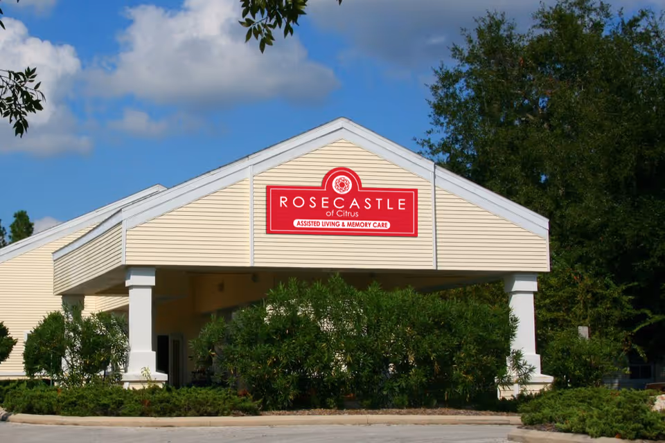 Front entrance of a single-story senior living building with a covered porte-cochere, landscaping, and a red 'Rosecastle' sign.