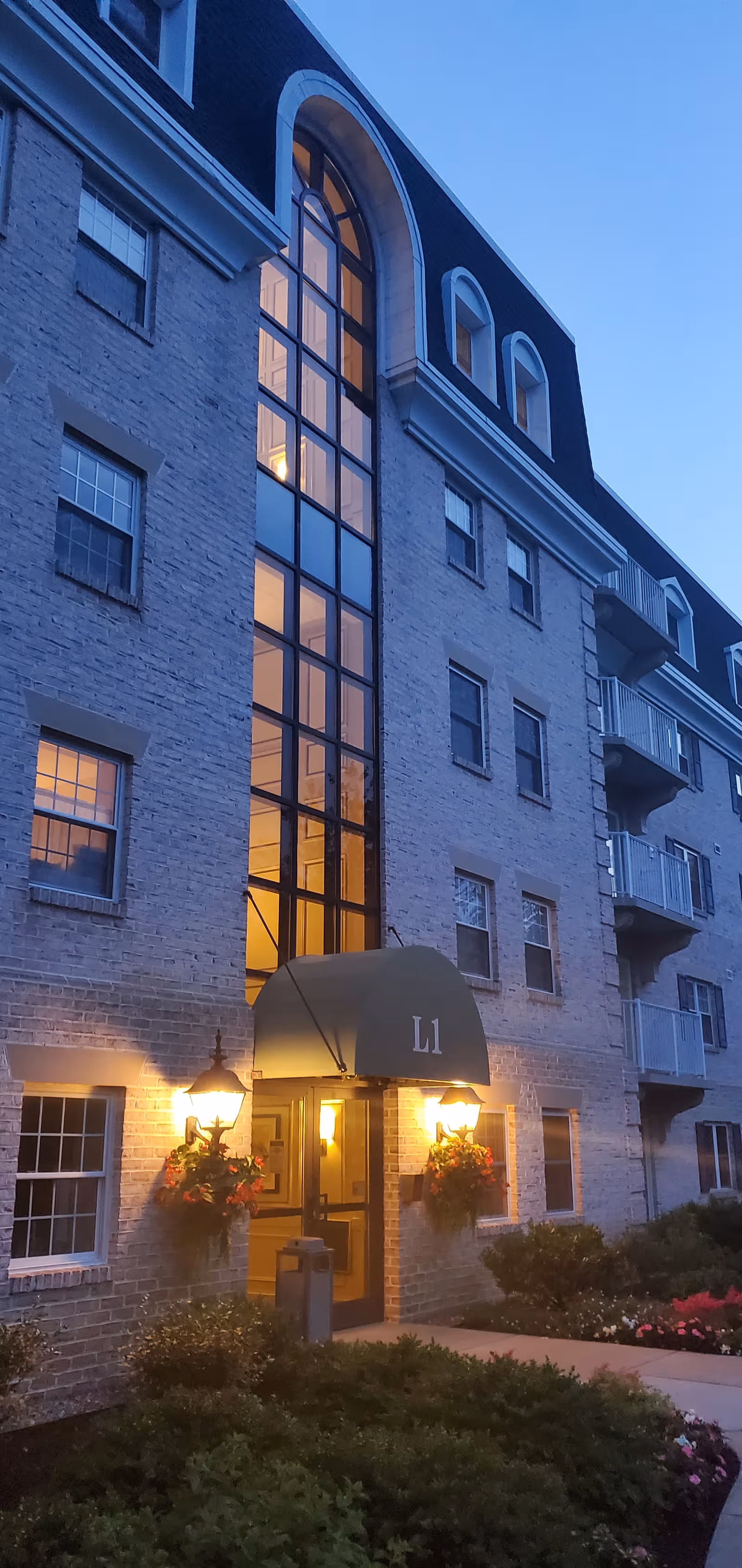 Exterior brick building with a lit entrance, arched window, and hanging flower baskets.