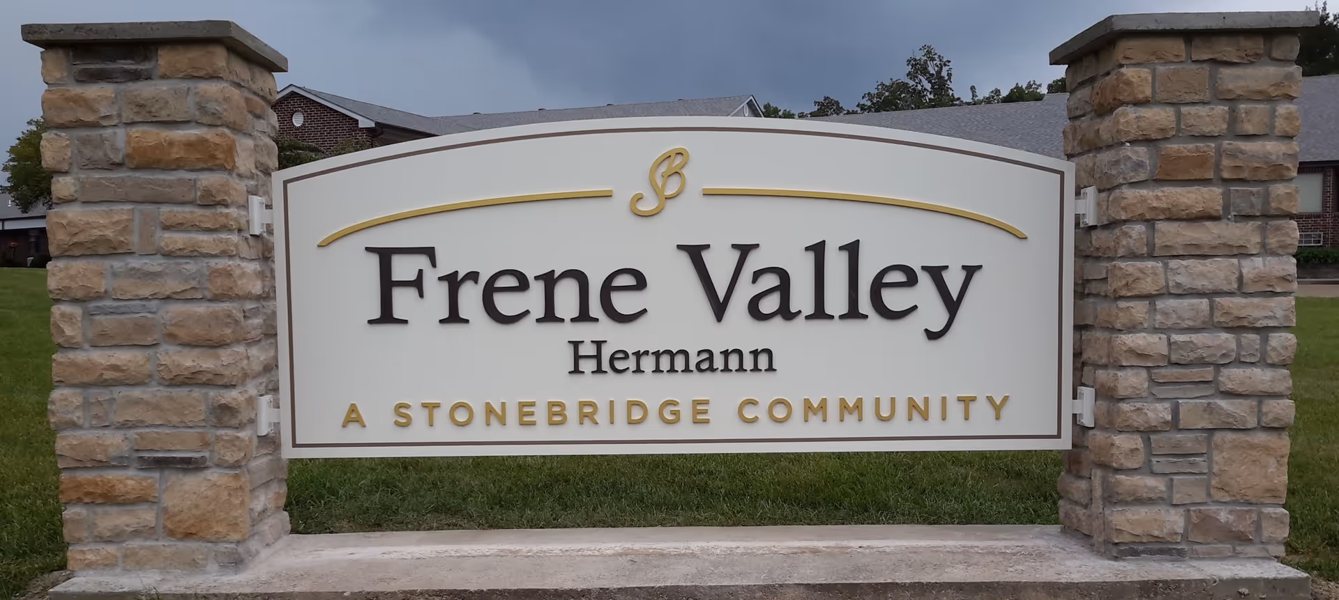 Stone-pillared entrance sign reading "Frene Valley Hermann A Stonebridge Community" with a lawn and building in the background.