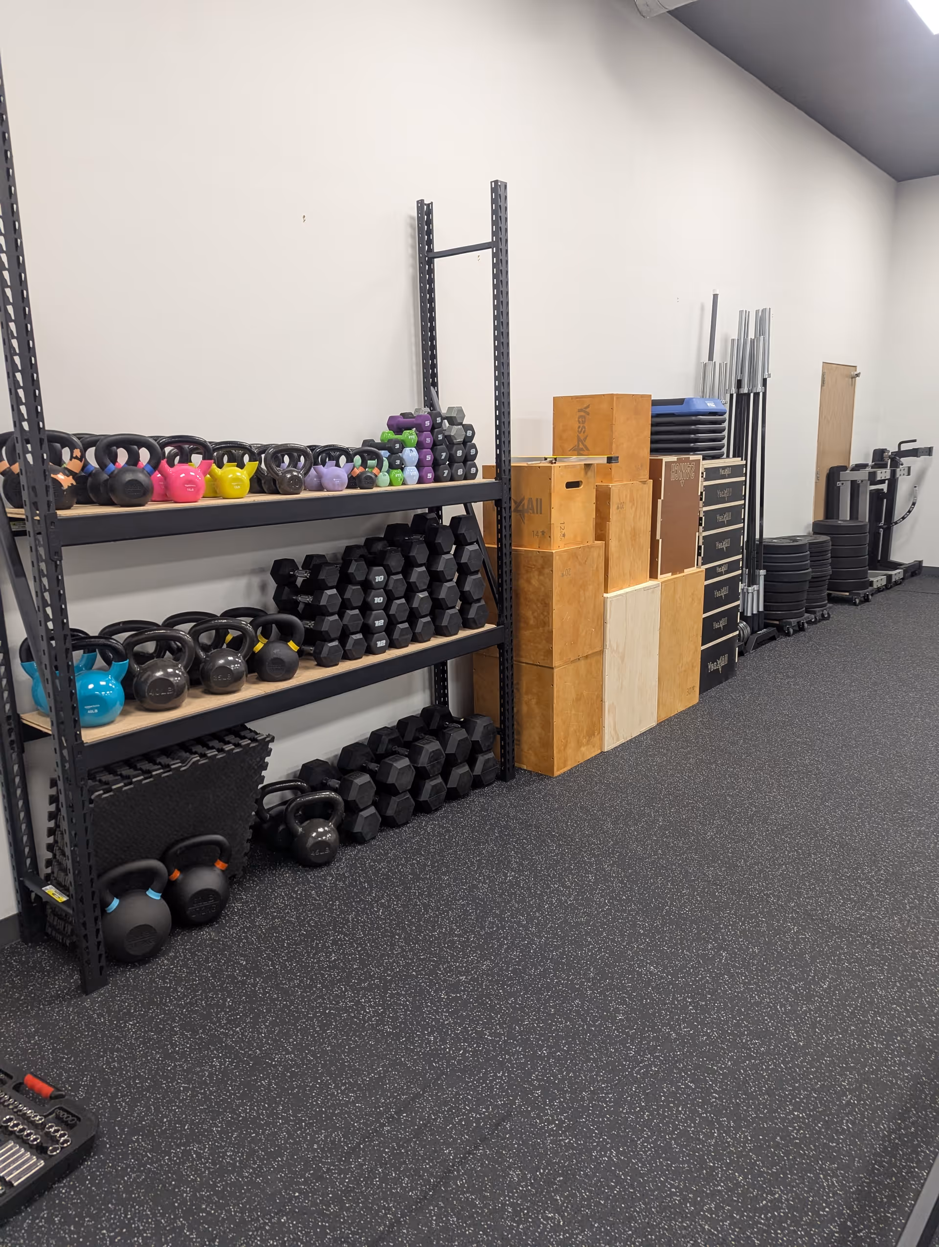 Industrial gym area with kettlebells, dumbbells, boxes, and barbells along storage racks.