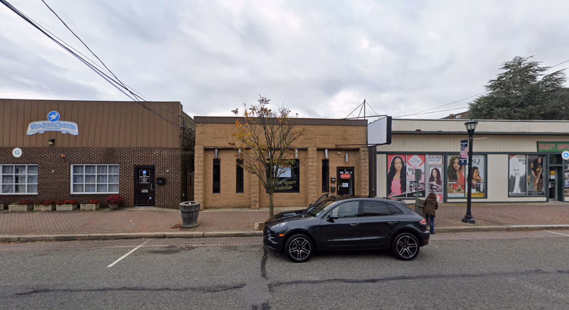 Row of small storefronts along a sidewalk with a black SUV parked in front.