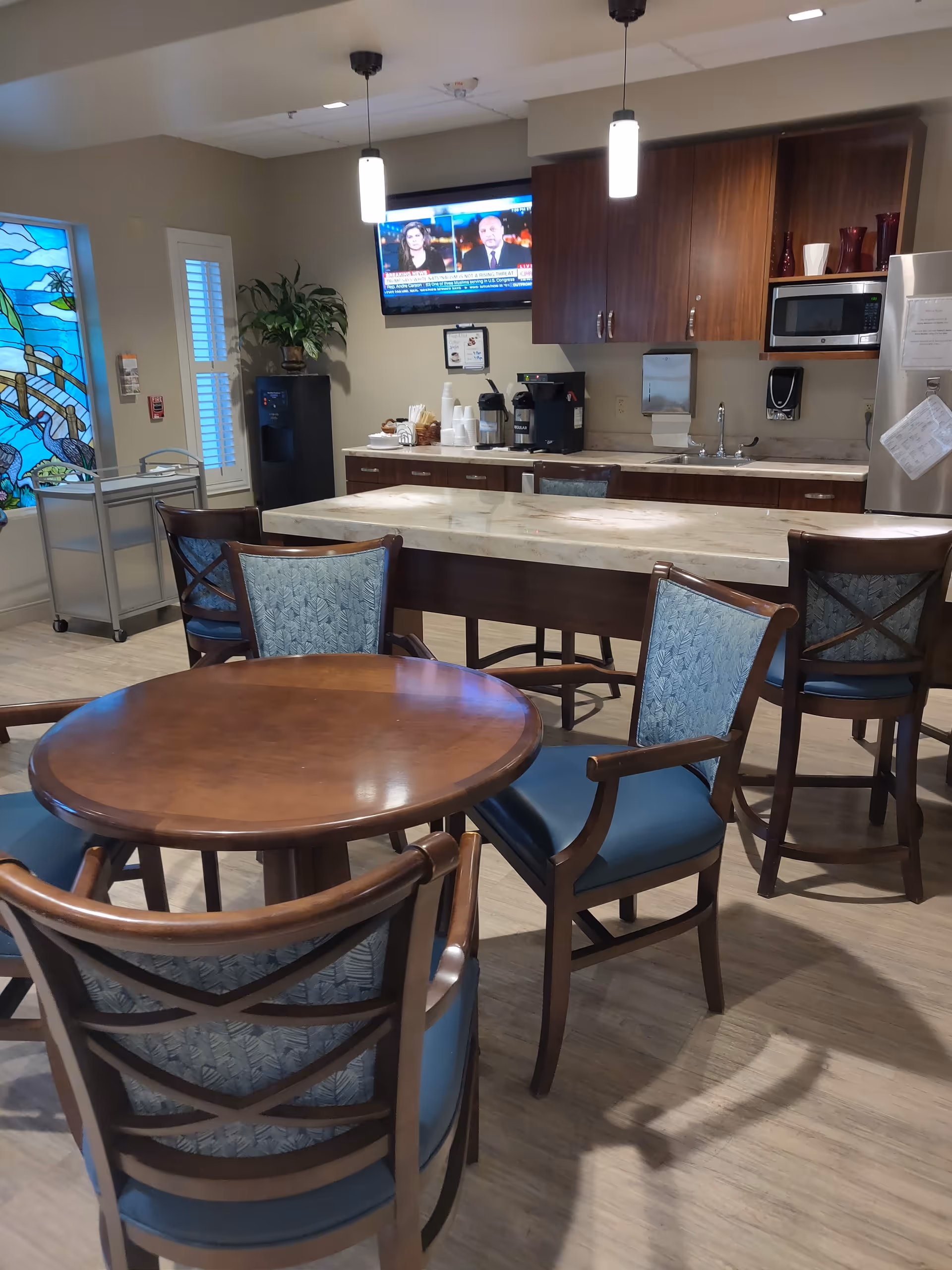Cozy communal dining area with round table, blue upholstered chairs, and kitchenette in background.